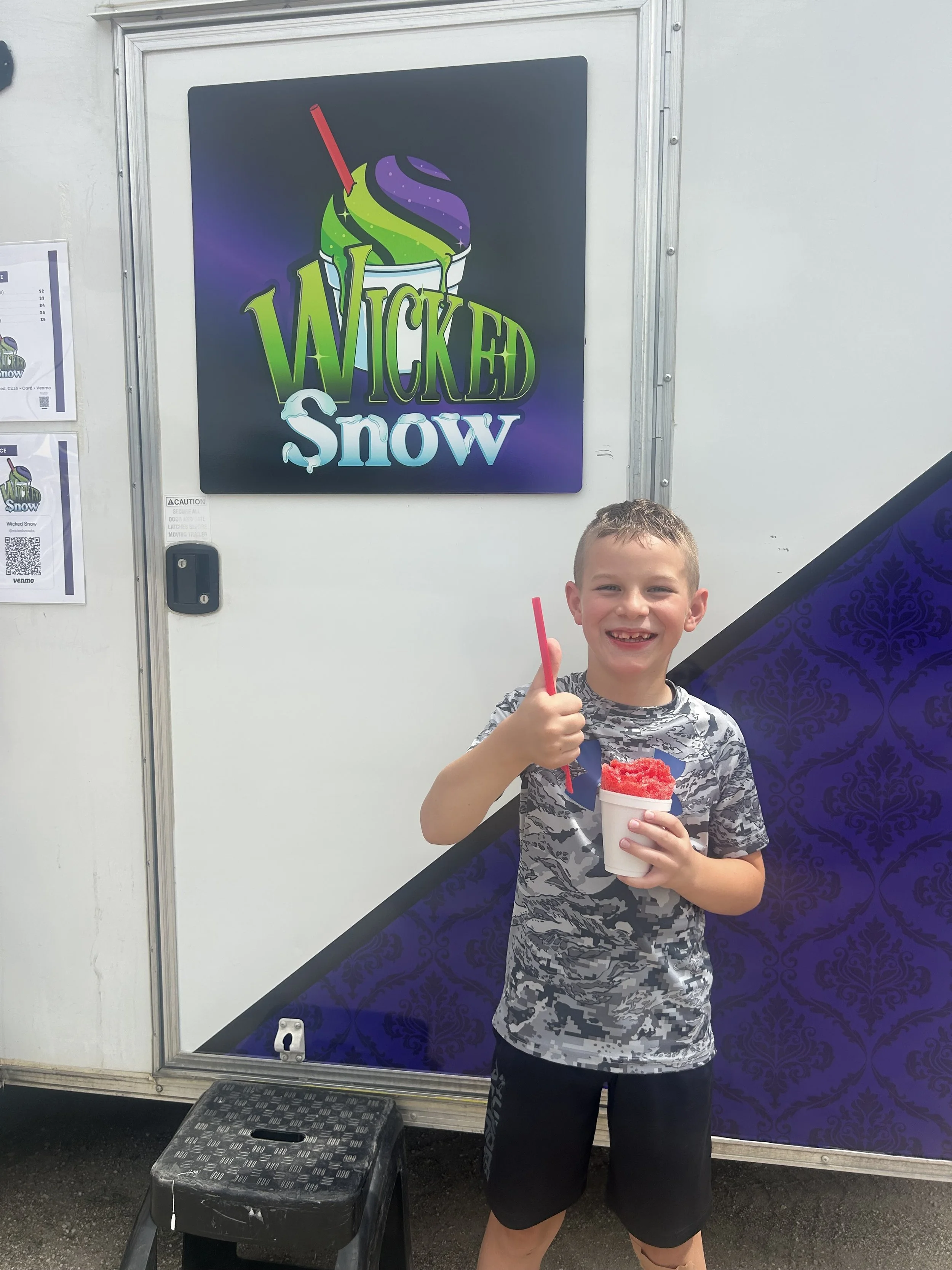 A young boy smiling and giving a thumbs up while holding a cup of red snow cone, standing in front of a food truck with a sign that says 'Wicked Snow.'