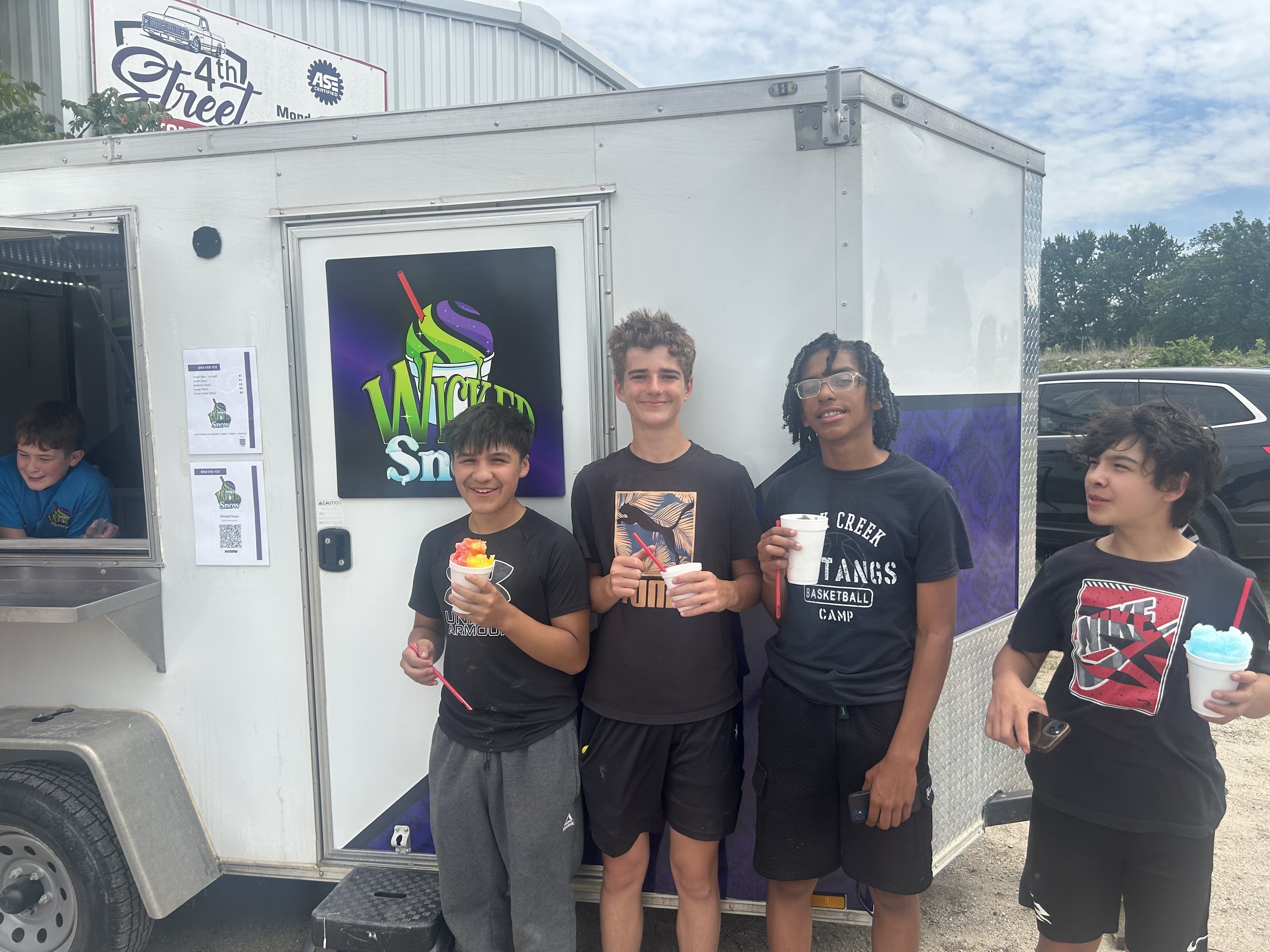 Four young people standing outside of a white ice cream truck holding cups of colorful shaved ice. The truck has a logo with a swirl of purple, green, and white ice cream with a red straw and the partial name 'Wicked Snow.' They are smiling and appear to be enjoying their treats.