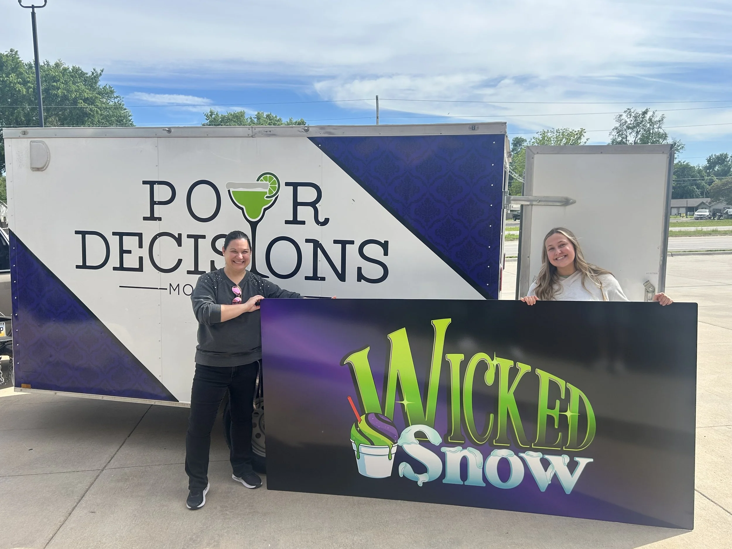 Two women standing outside near a food truck labeled 'Pour Decisions' and a colorful sign that reads 'Wicked'. One woman has dark hair, wearing a black shirt with pink sunglasses hanging on her collar. The other has long blonde hair, wearing a light-colored shirt. Blue sky and trees can be seen in the background.