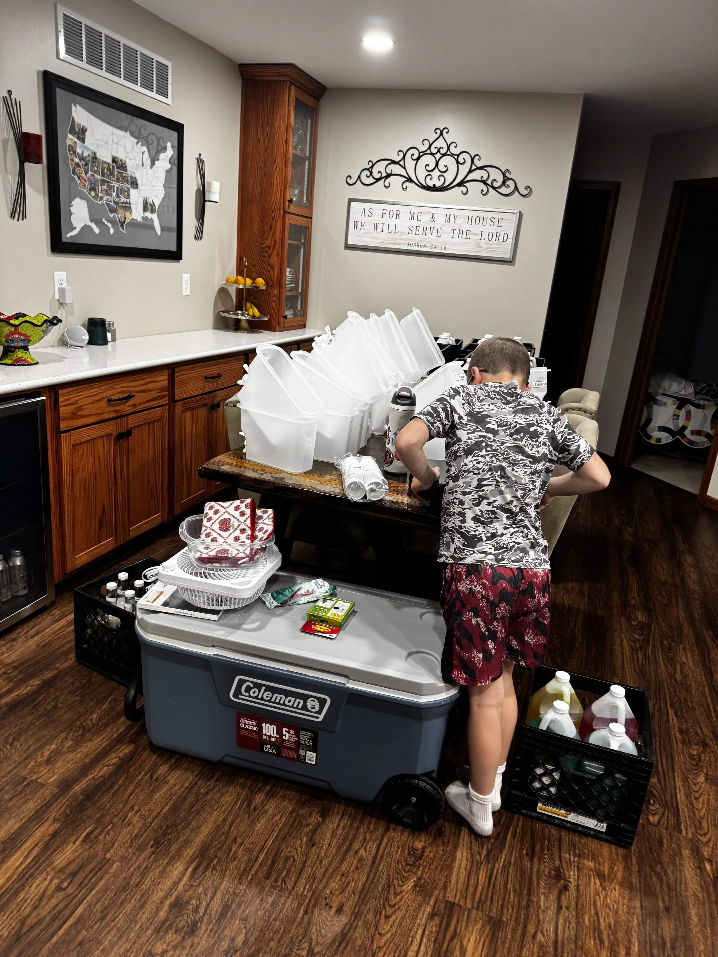 A boy in pajamas standing in a kitchen, organizing plastic containers on a table, with a large Coleman cooler nearby and kitchen cabinets in the background.