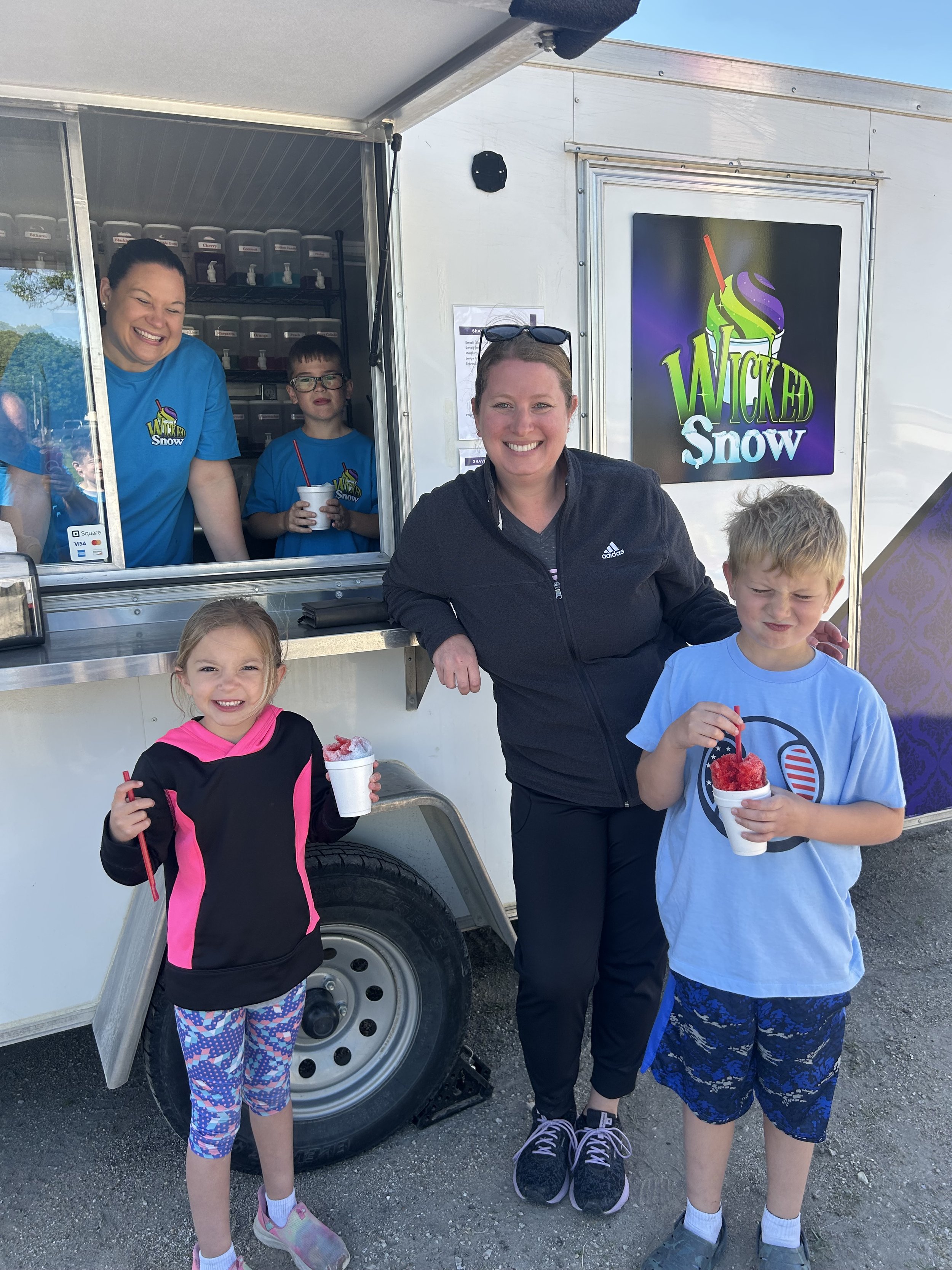 A group of people, including children and adults, standing outside near a mobile ice cream or snow cone stand. The children are holding cups of shaved ice with red syrup. The stand has a large sign that says 'Wicked Snow' with bright colors and a picture of a snow cone on it. The people are smiling and enjoying their treats.