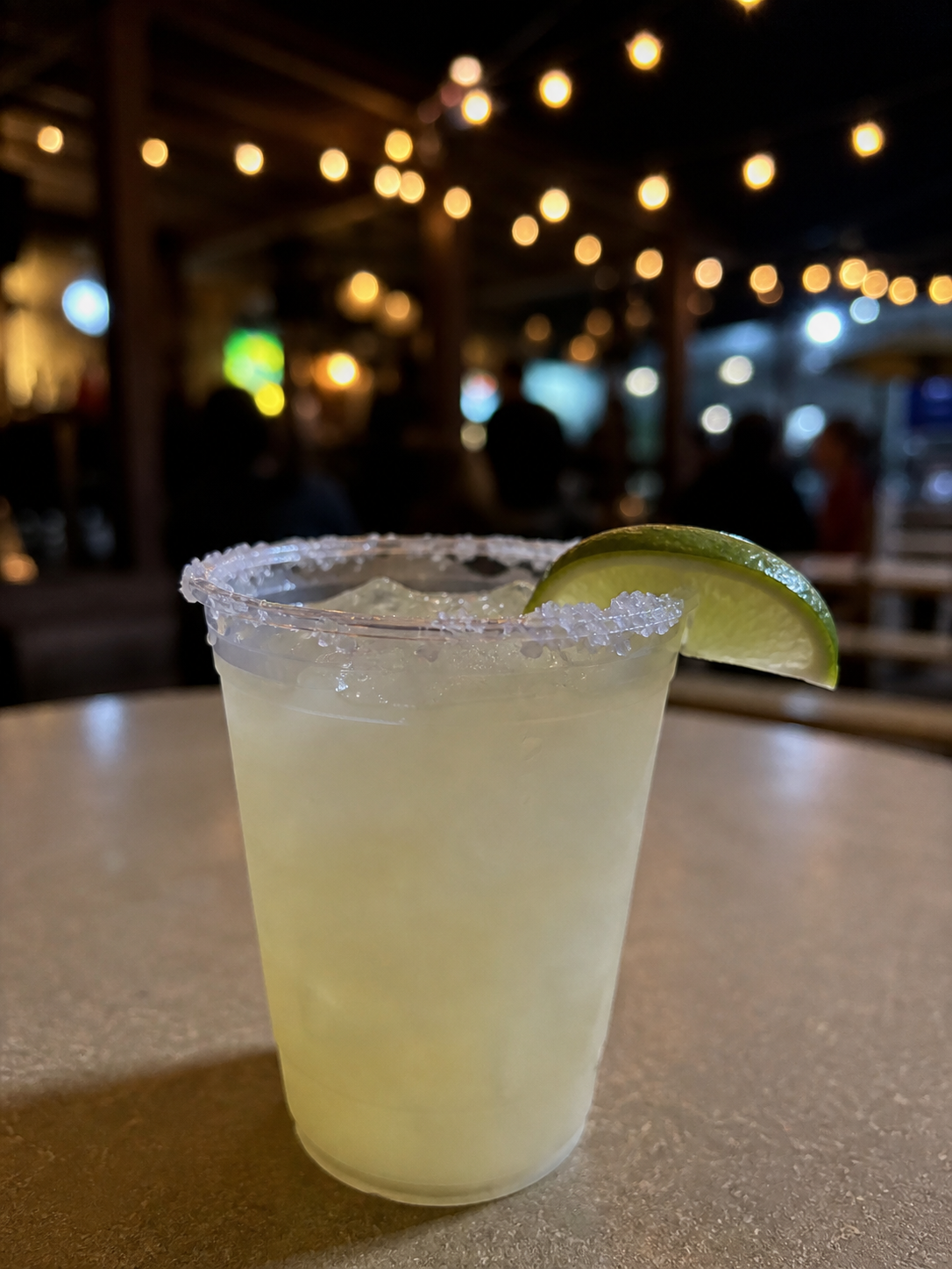 A margarita cocktail with a salted rim and a lime wedge garnish, on a table in a dimly lit bar or restaurant with hanging string lights and blurred patrons in the background.