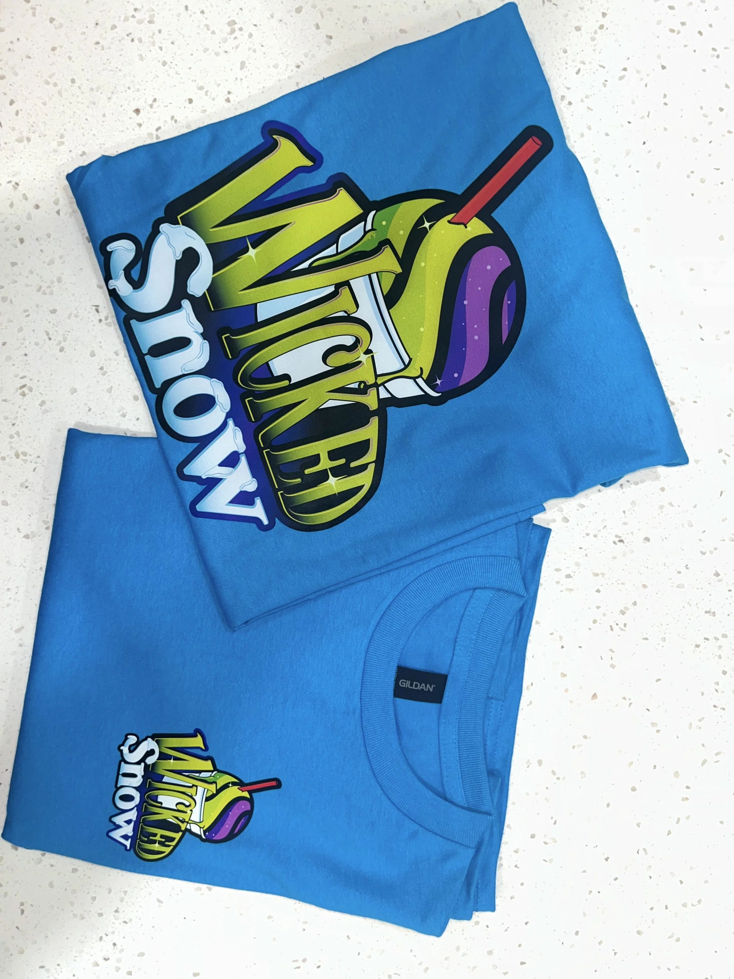Blue T-shirt with a colorful graphic design, featuring a stylized cocktail glass with a purple liquid, yellow lemon wedge, green lime, red straw, and the words "Never Stop" in bold letters.