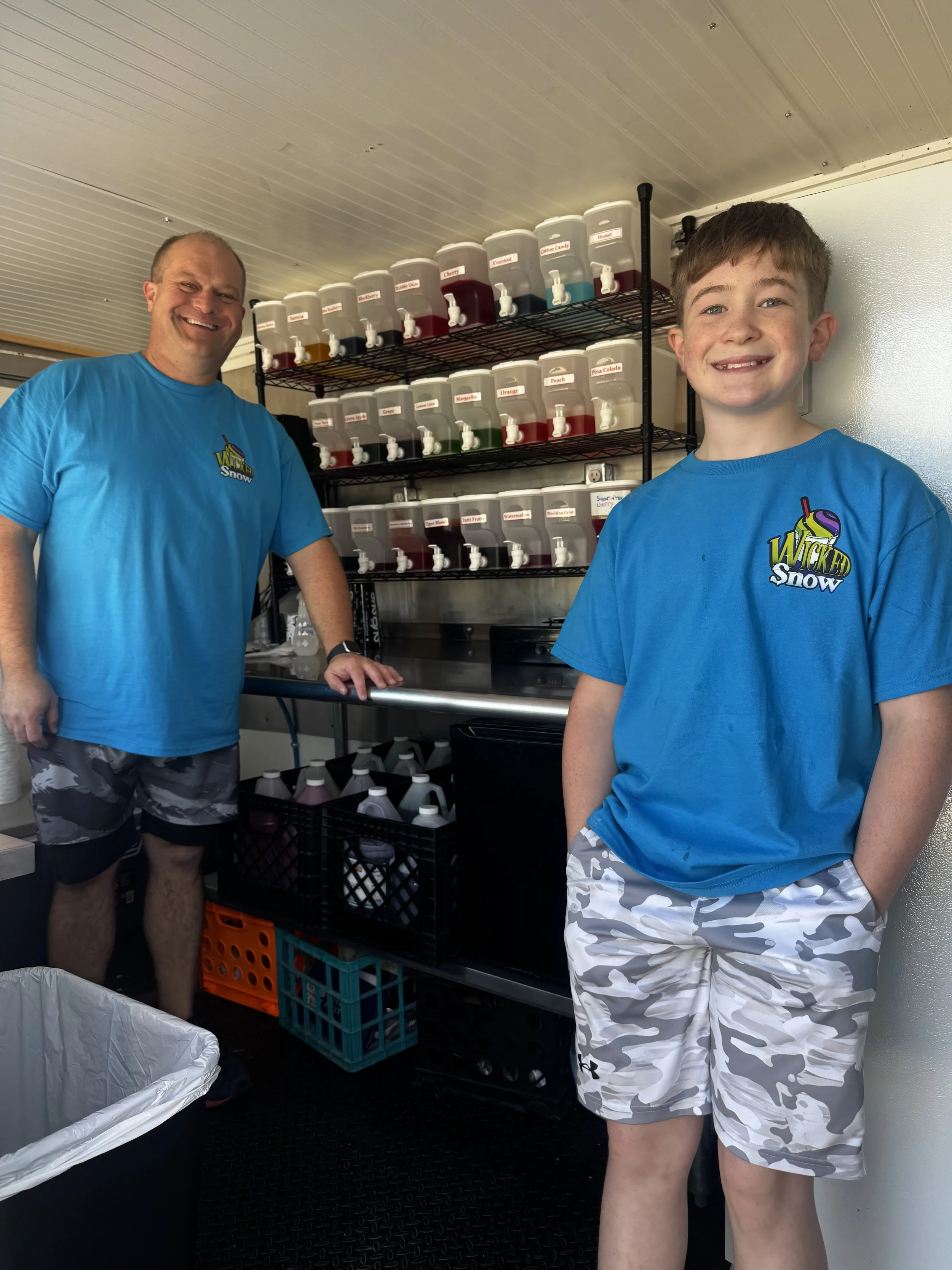 Two smiling males in blue shirts. The man on the left is bald and wearing camouflage shorts and a watch, standing with one hand on a metal counter. The boy on the right has short brown hair, is wearing a blue T-shirt with a colorful logo, and white shorts with gray camouflage pattern. Behind them, there is a shelf with multiple clear dispensers filled with colorful liquids with labels, possibly for a snow-cone or ice treat stand. There is also a laundry basket in the foreground and a white wall on the right.