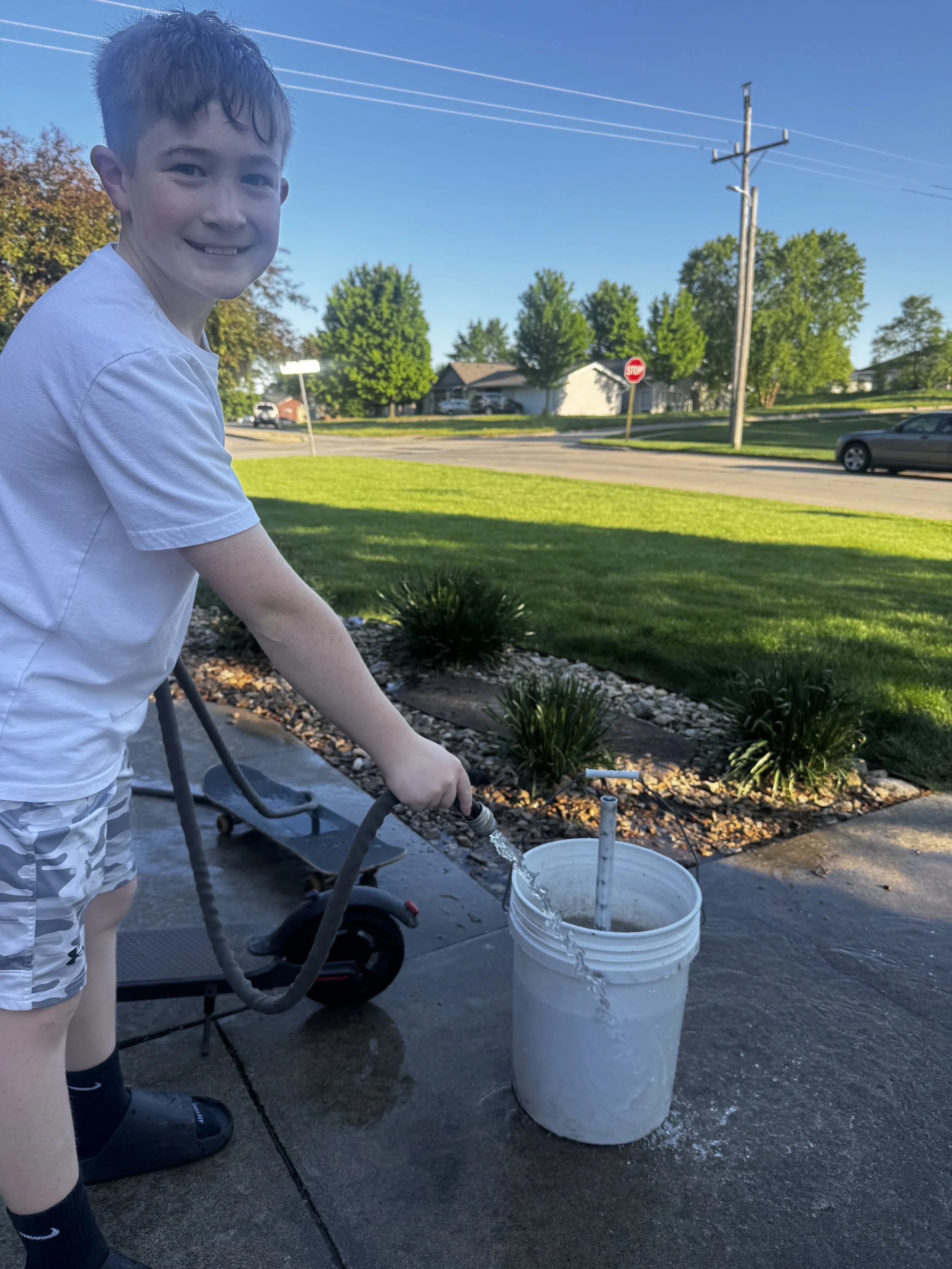 A young boy wearing a white t-shirt, camouflage shorts, and black shoes is watering a garden with a garden hose. The hose is positioned over a white bucket, which is placed on a concrete driveway. Behind him, there is a lawn, some bushes, and a street with a stop sign, a parked car, and trees under a clear blue sky.