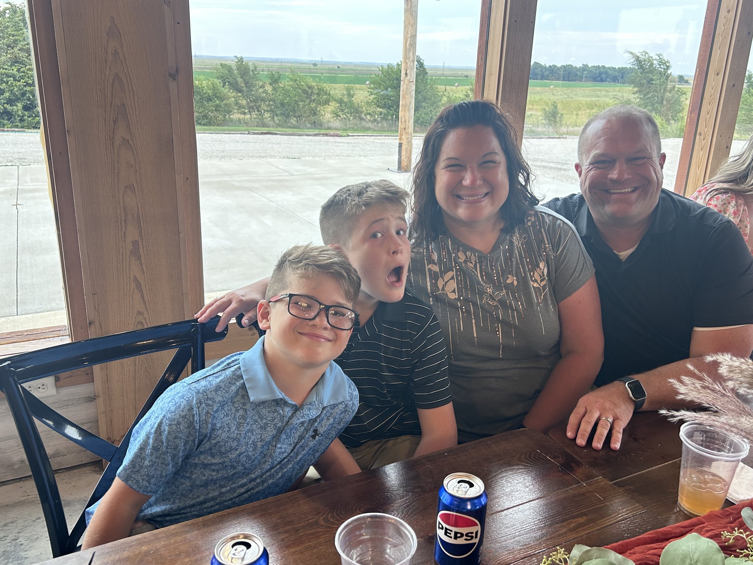 A group of five people smiling and posing inside a rustic building with large windows overlooking a rural landscape. There are drinks and food on the wooden table in front of them.