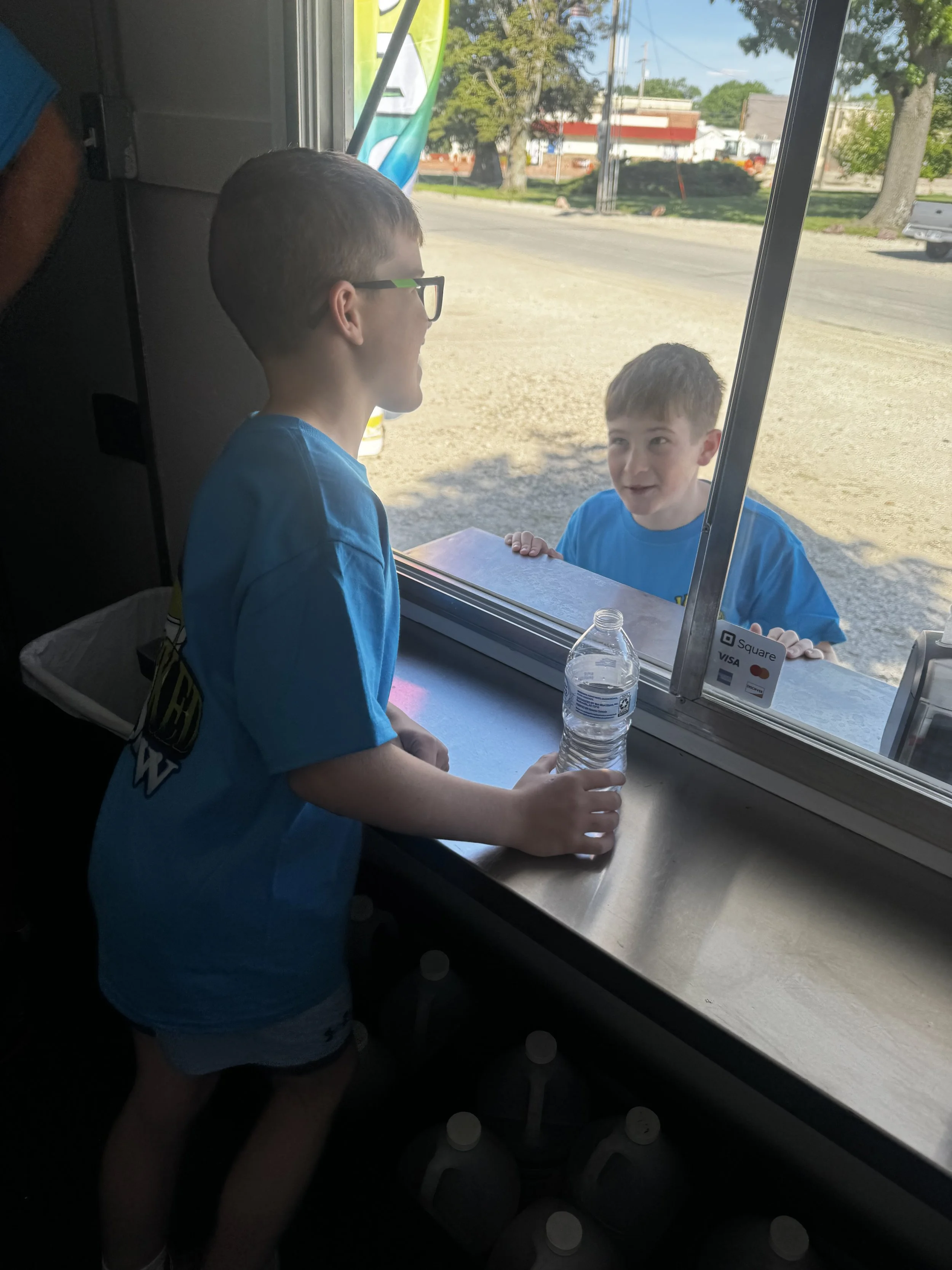 Two young boys, one wearing glasses, are at a window counter, with one boy inside and the other outside, exchanging or handing over a water bottle. The scene is outside on a sunny day, with trees and buildings visible through the window.