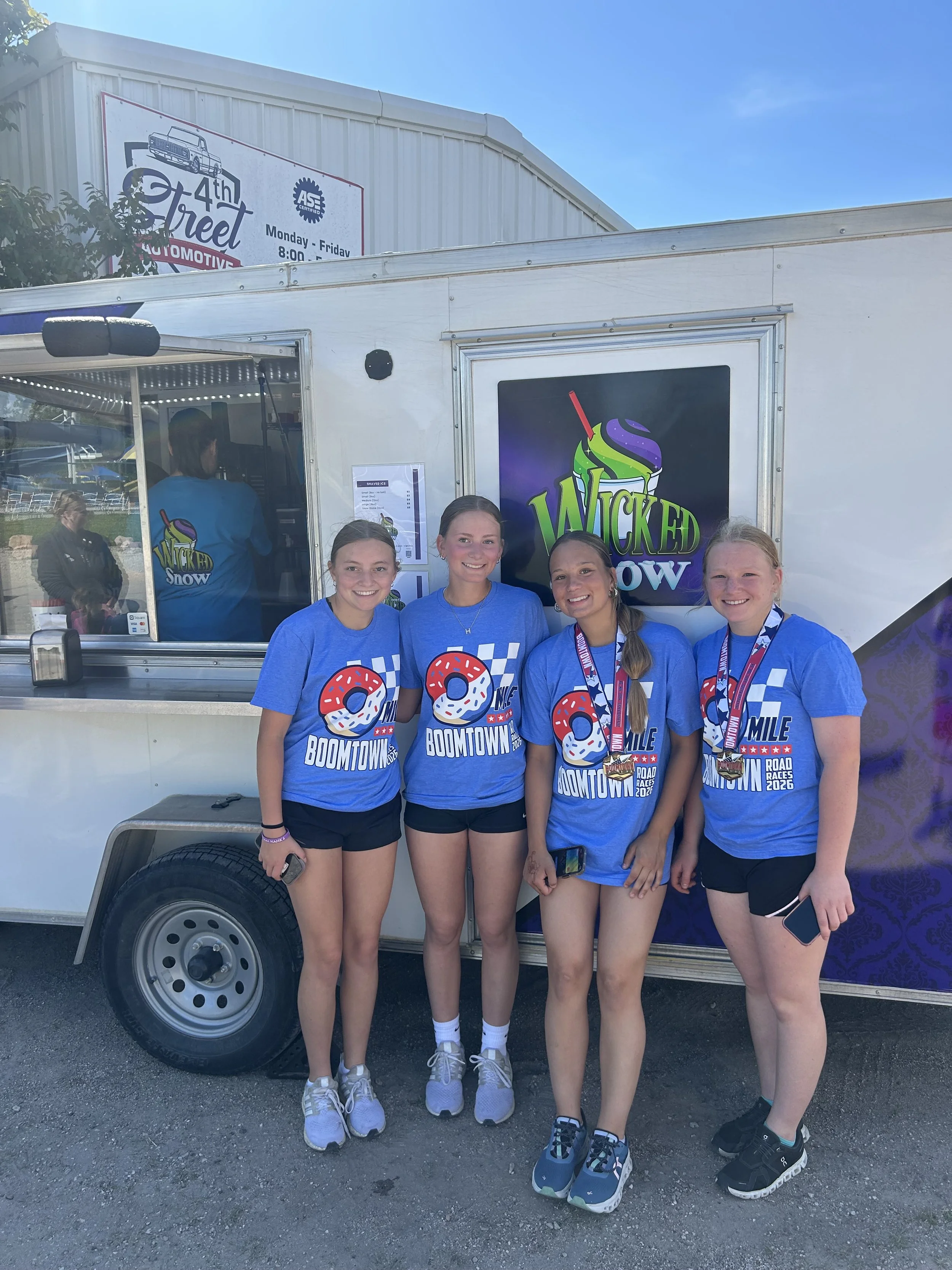 Four young women wearing matching blue t-shirts with a donut graphic and the words "BOOMTOWN" and "MILE" are standing in front of a white food truck with a large sign that reads "WICKED SHOW." They appear to be at an outdoor event, smiling and posing for the photo, with medals around their necks.
