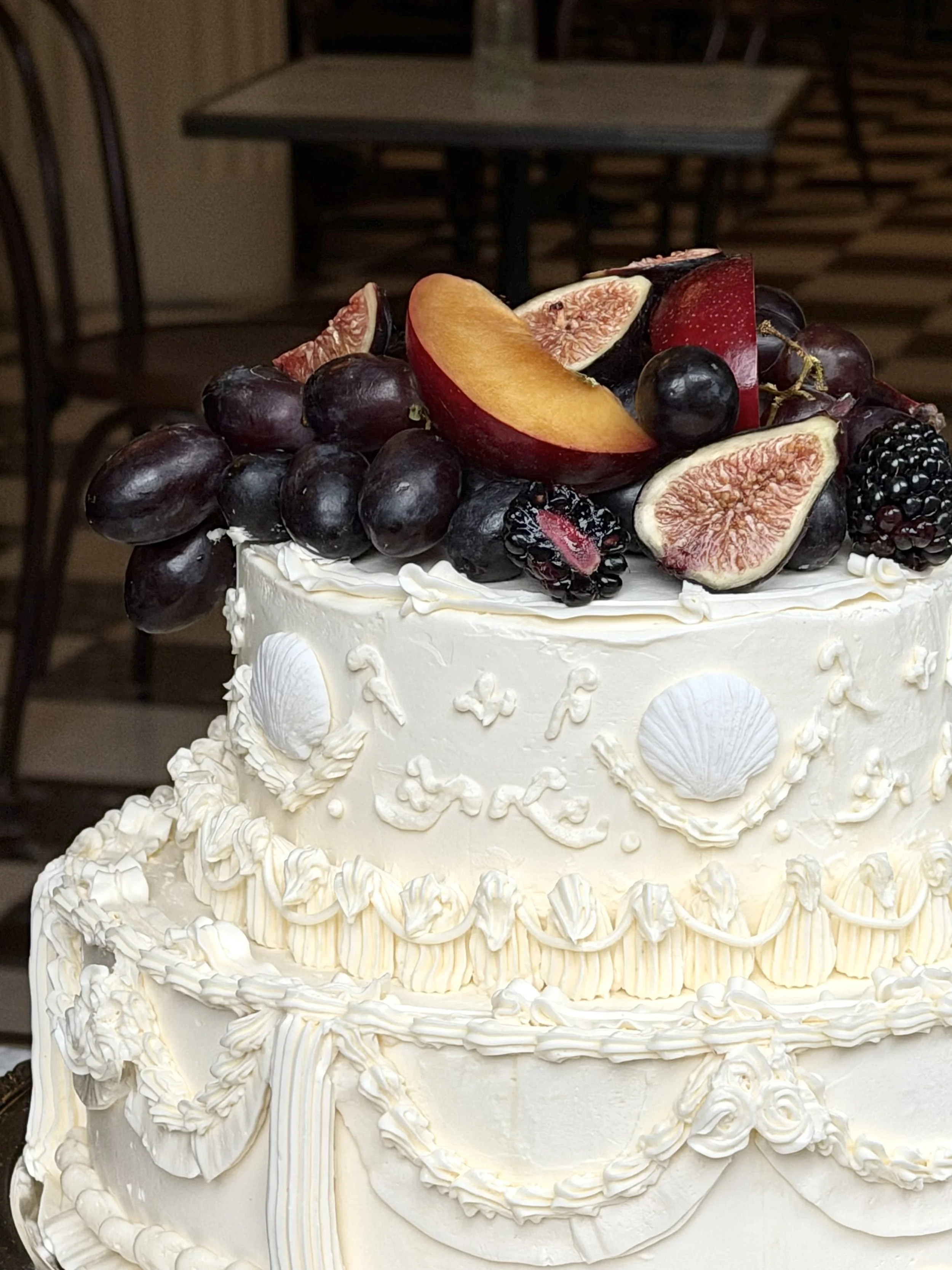 A white wedding cake decorated with piped white icing, featuring fresh fruit including grapes, figs, blackberries, and a slice of peach on top.