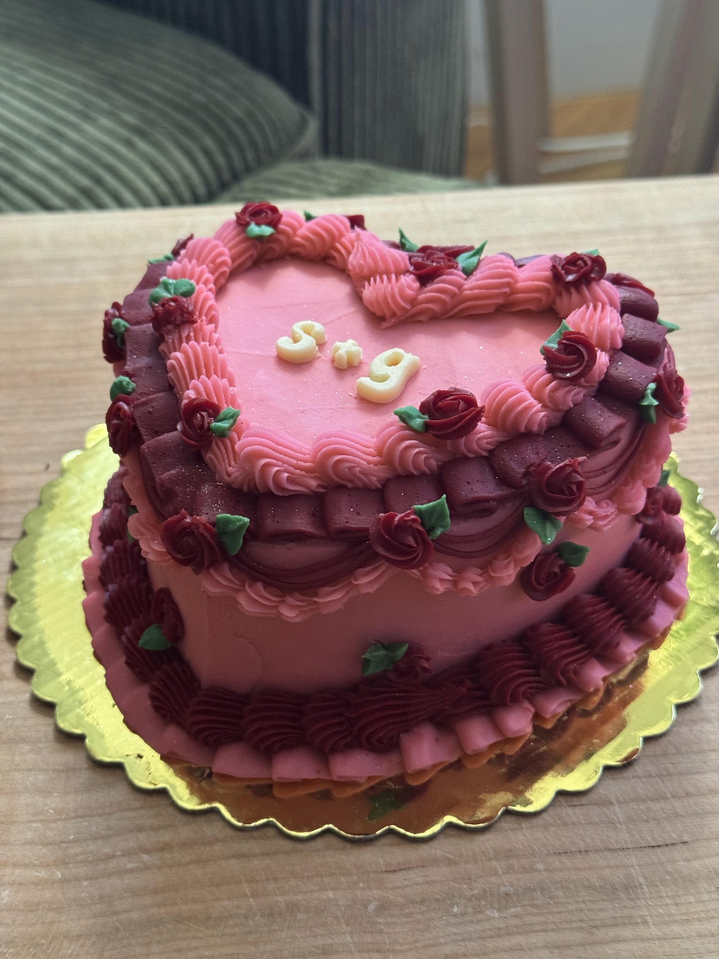A round pink and dark red heart-shaped cake decorated with roses and green leaves, with the name 'George' written on top in yellow.