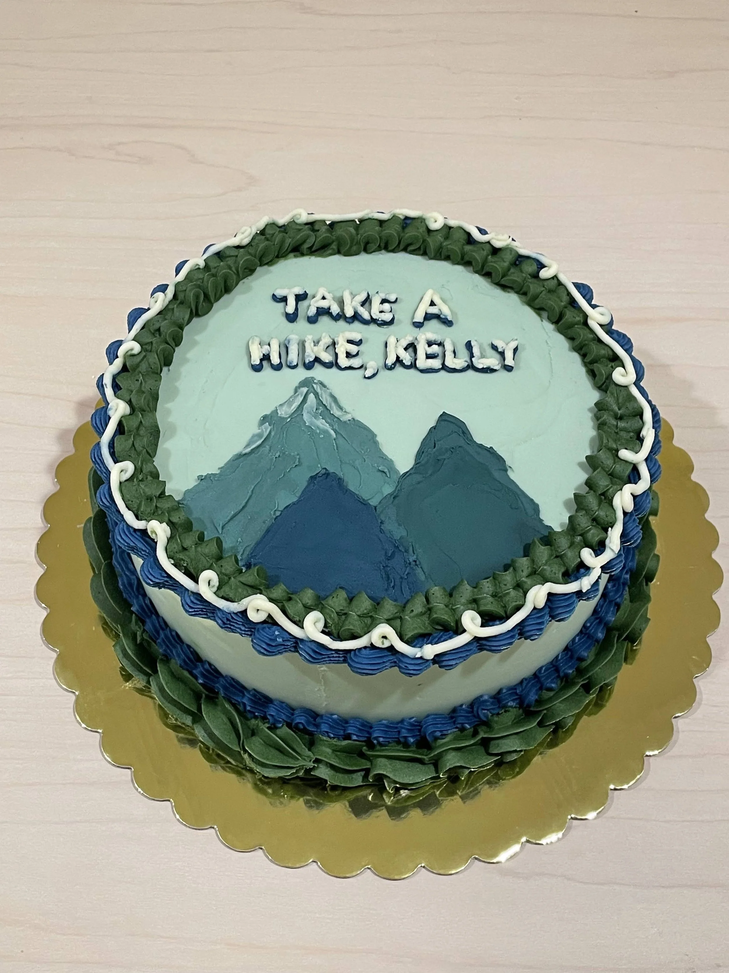 A round cake decorated with mountain scenery and the message "Take a Hike, Kelly" written on top, with green, blue, and white frosting.