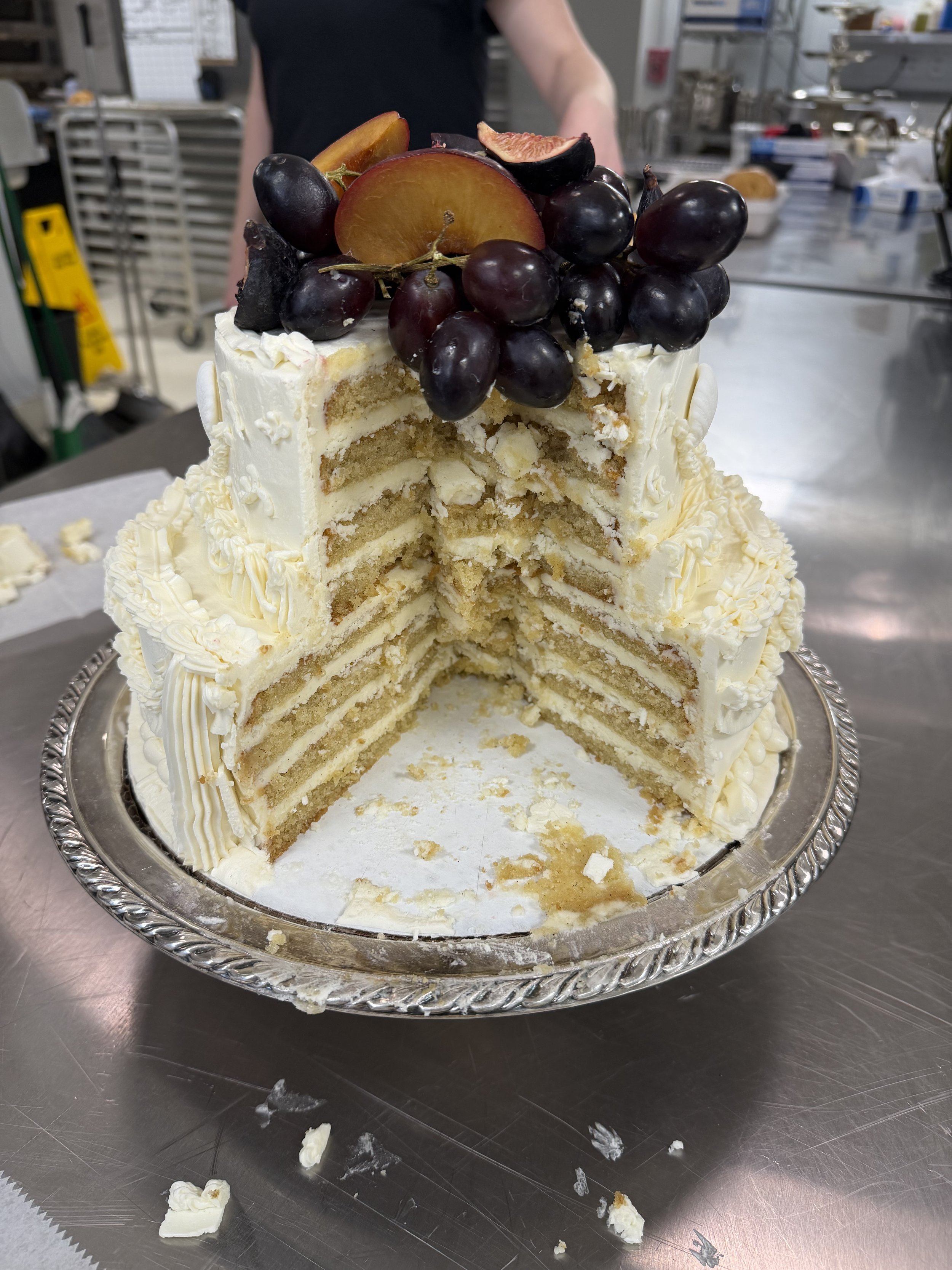 A large, partially eaten layered vanilla cake with white icing, topped with black grapes, a peach slice, and a fig, on a silver tray, on a stainless steel table in a kitchen or bakery setting.