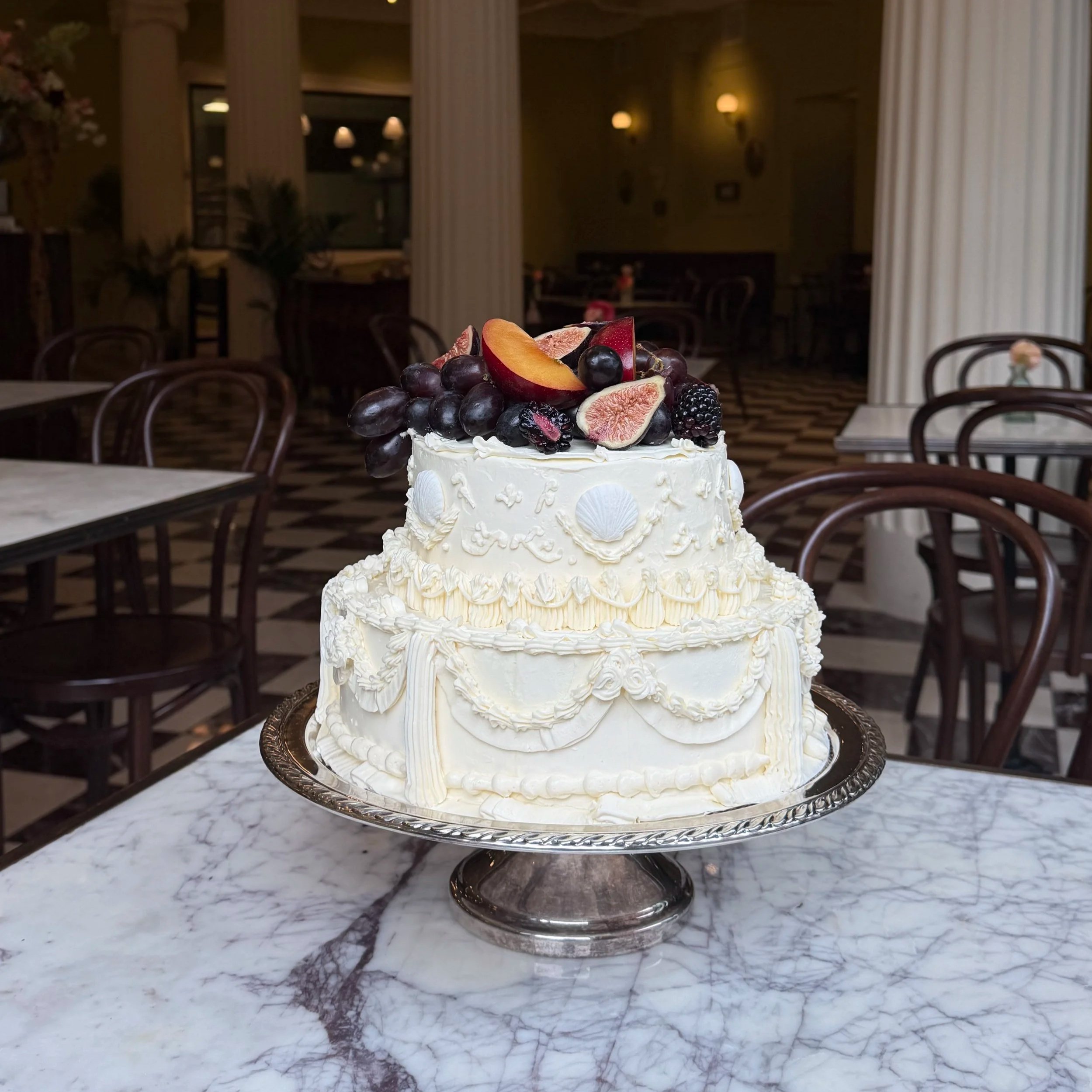 A white wedding cake decorated with fresh berries and sliced figs on top, placed on a silver cake stand on a marble table in a dimly lit restaurant or banquet hall with wooden chairs and tables in the background.
