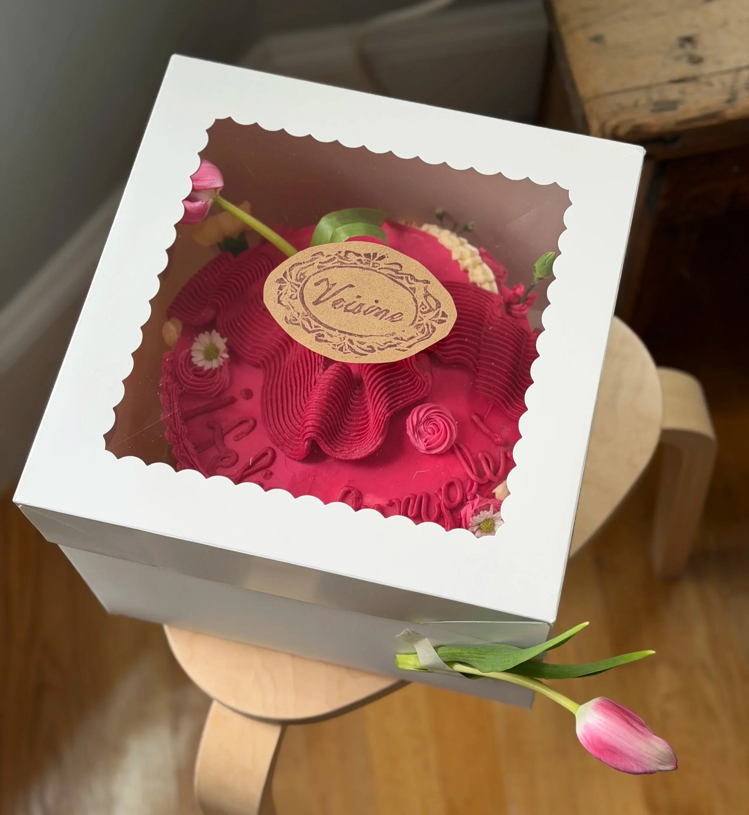 A Valentine's Day cake box with a pink flower and green leaves sticking out at the side, showing part of a pink cake inside. The box has a transparent window and a label that reads 'Valentine'.