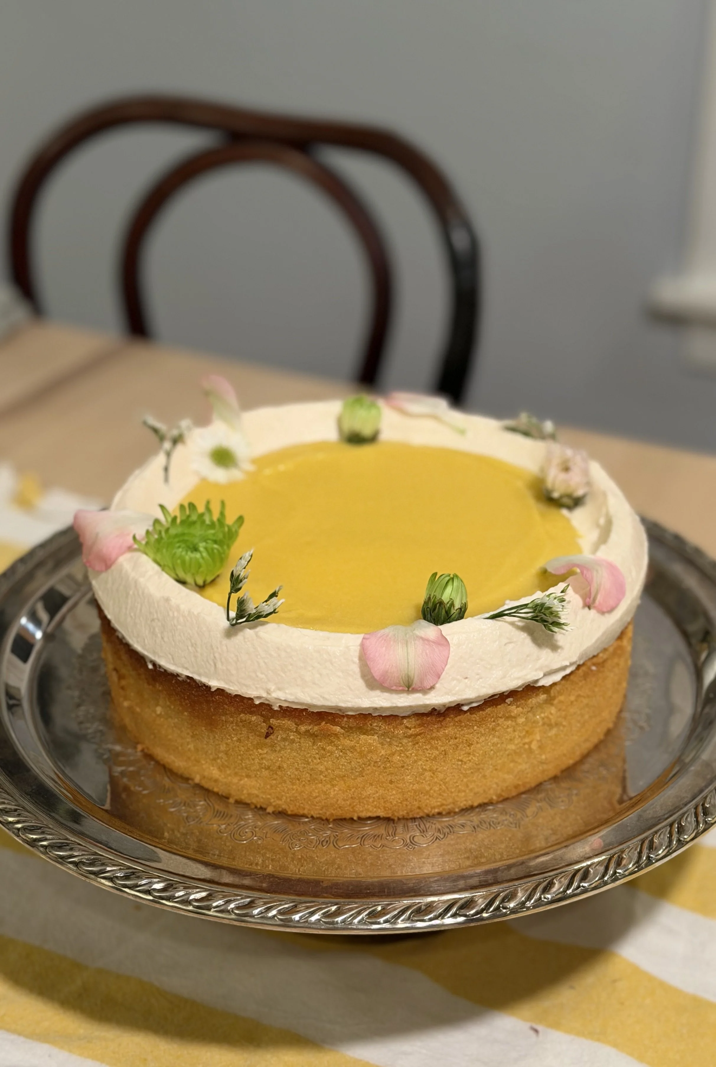 A round cake with yellow filling topped with edible flowers and a cream border, placed on a silver tray.