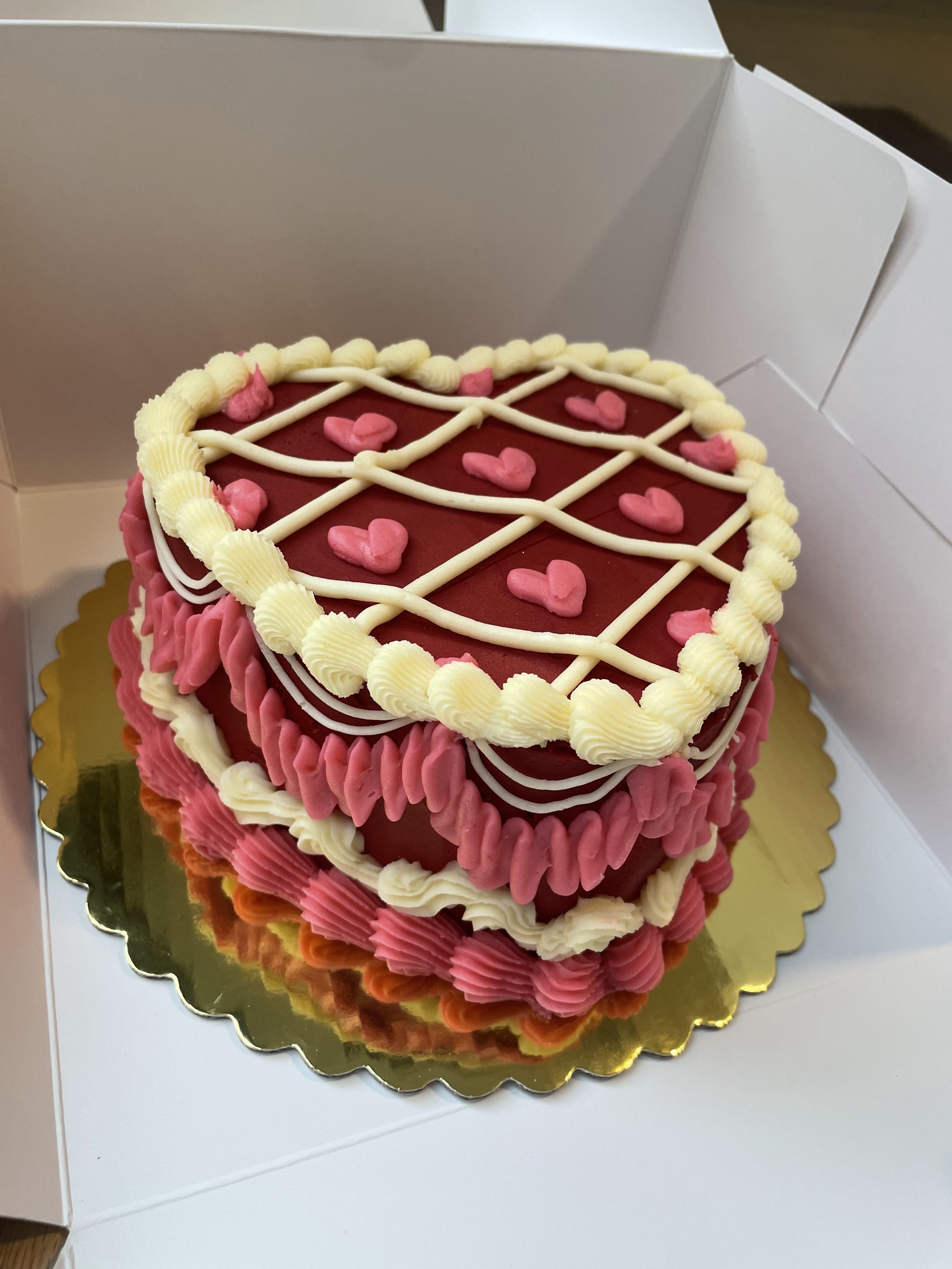 Heart-shaped cake with red, pink, and white frosting, decorated with small pink hearts and white icing lines, in a white box.