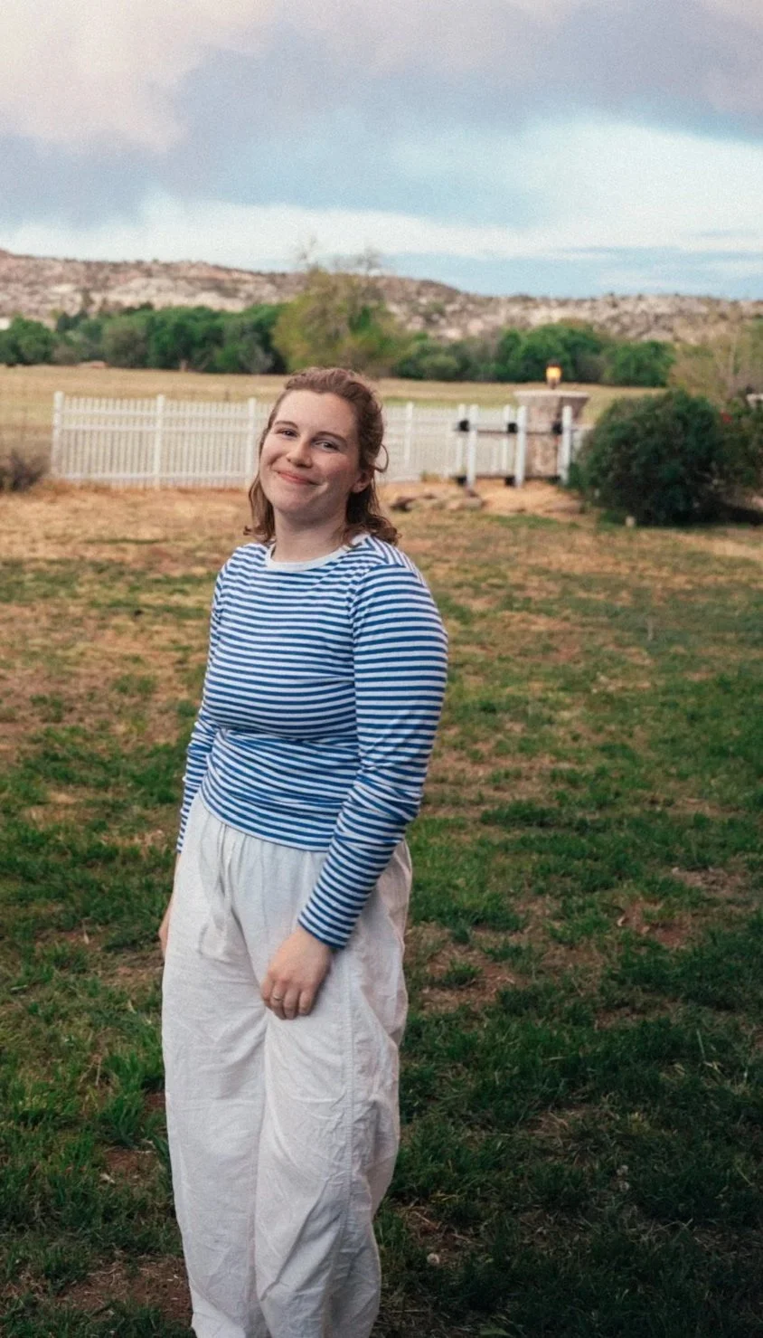 A young woman with curly hair smiling outdoors in a grassy backyard, wearing a blue and white striped long-sleeve shirt and white pants, with a fence, trees, and hills in the background.