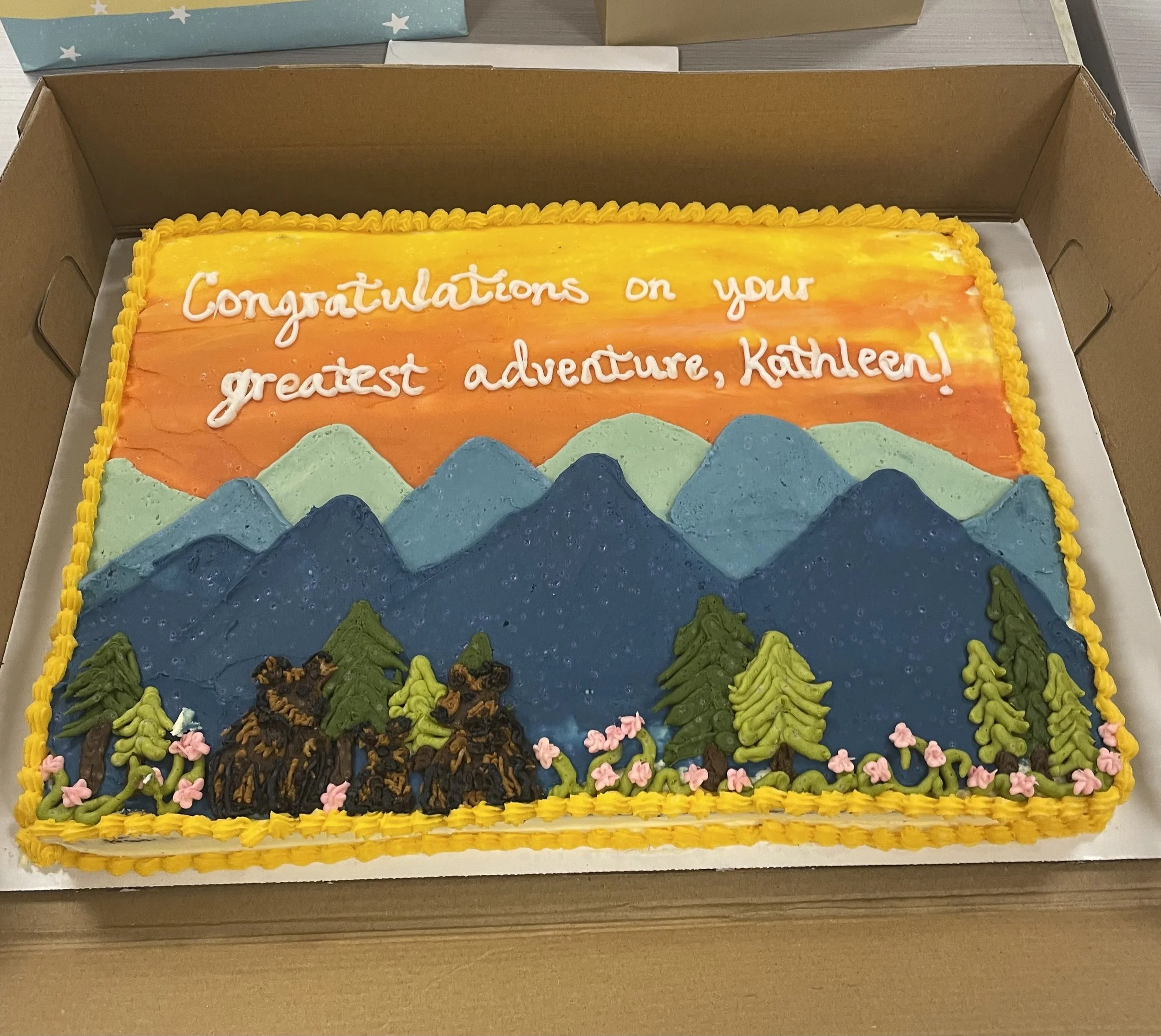 A colorful rectangular cake with icing that says 'Congratulations on your greatest adventure, Kathleen!' in white piping. The cake is decorated with mountain-like icing shapes in various shades of blue, green, and pink, and has a yellow piped border along the edges.