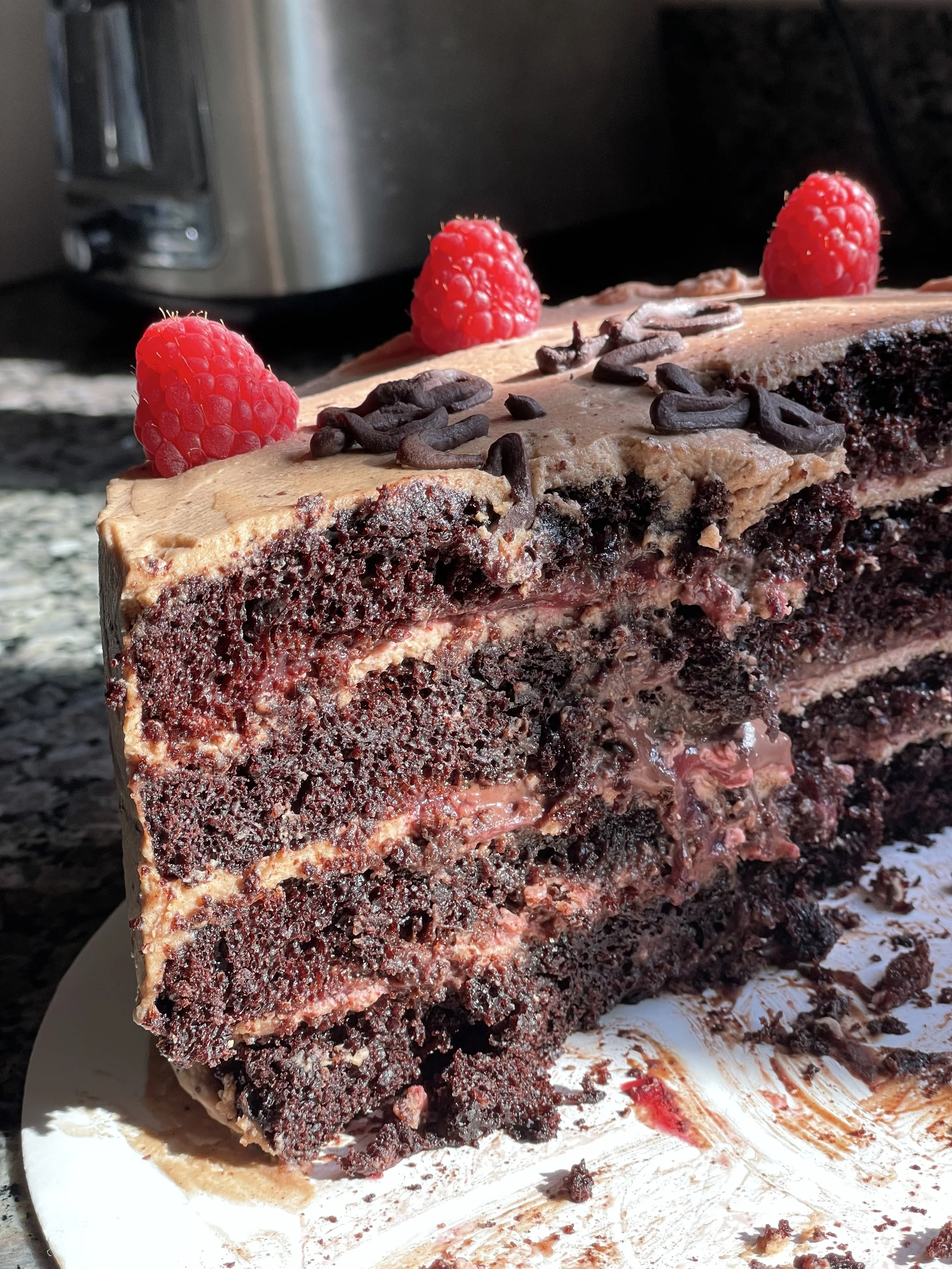 A sliced chocolate cake with raspberries and chocolate shavings on top, with some crumbs and frosting on a white plate.