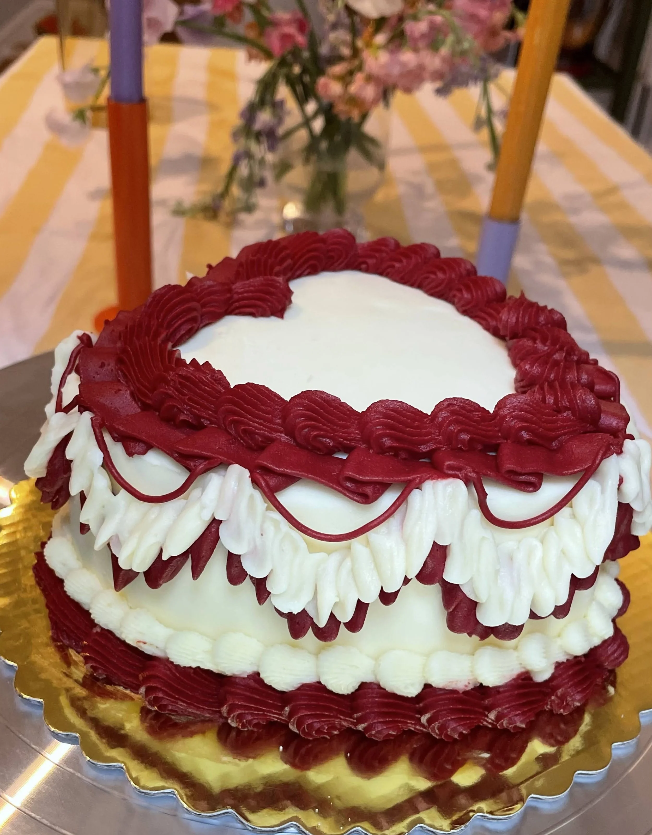 A birthday cake decorated with red and white piping, with a heart-shaped arrangement of red icing on top, and a floral arrangement in the background on a table with yellow and white striped tablecloth.