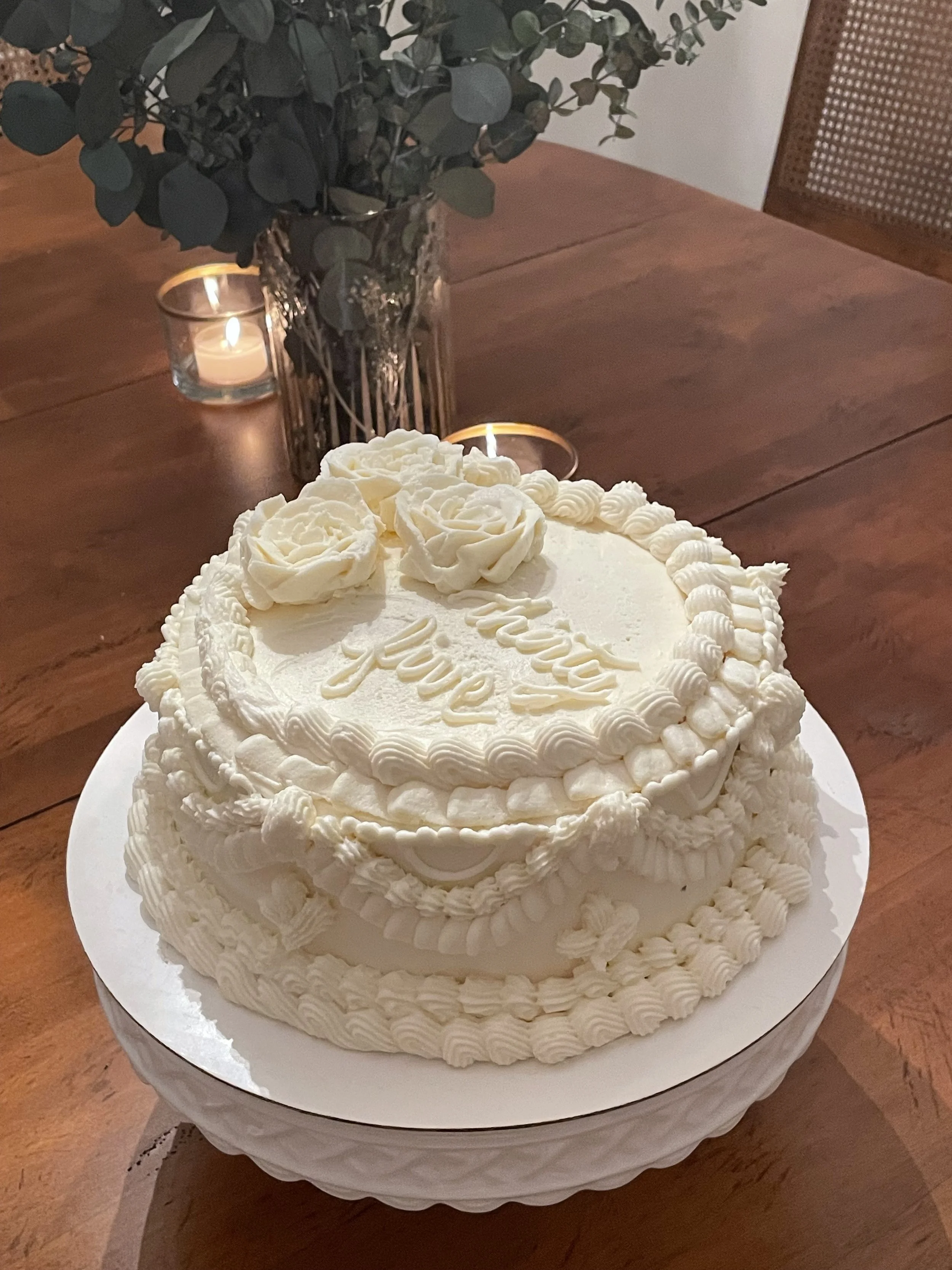 A white frosted birthday cake with floral decorations and the words 'Happy Birthday' written on top, placed on a round white stand. In the background, a glass vase with green leafy branches and two small candles in glass holders are visible on a wooden table.