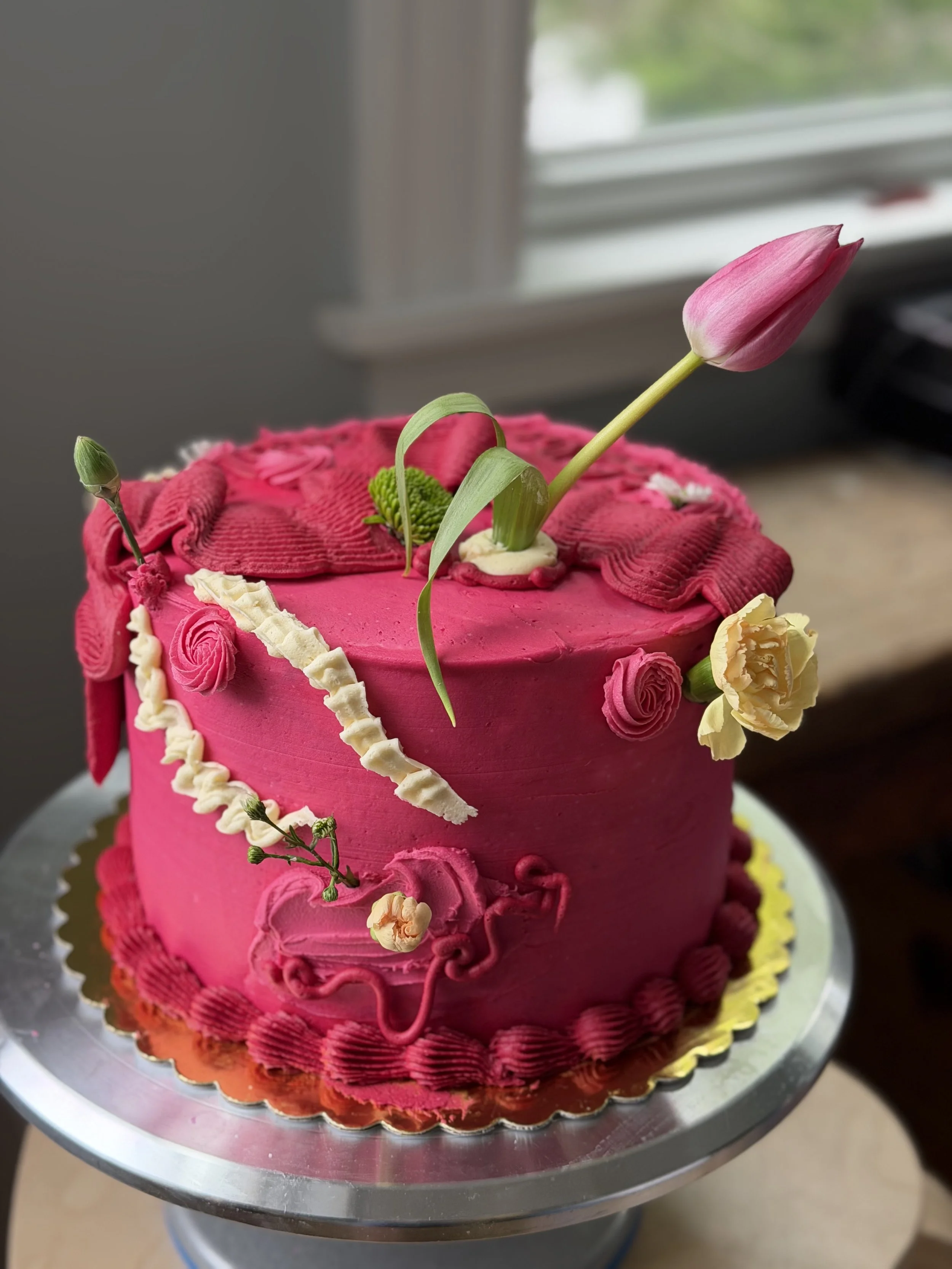 A round pink decorated cake with flowers and a pink tulip on top, displayed on a silver cake stand.