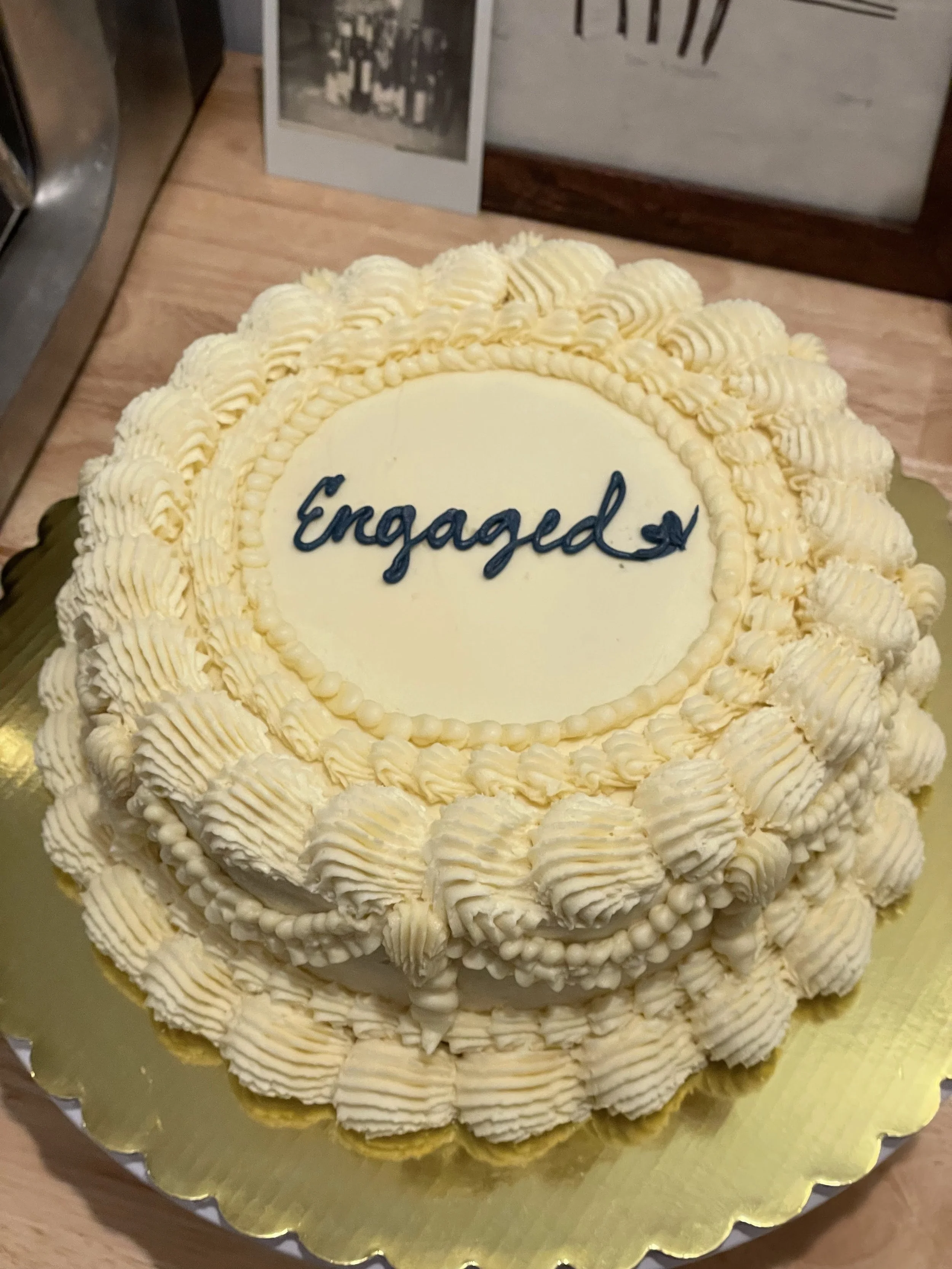 A decorated cake with the word "Engaged" written on it. The cake has a large number of white frosting rosettes and trim, and sits on a gold cake board.