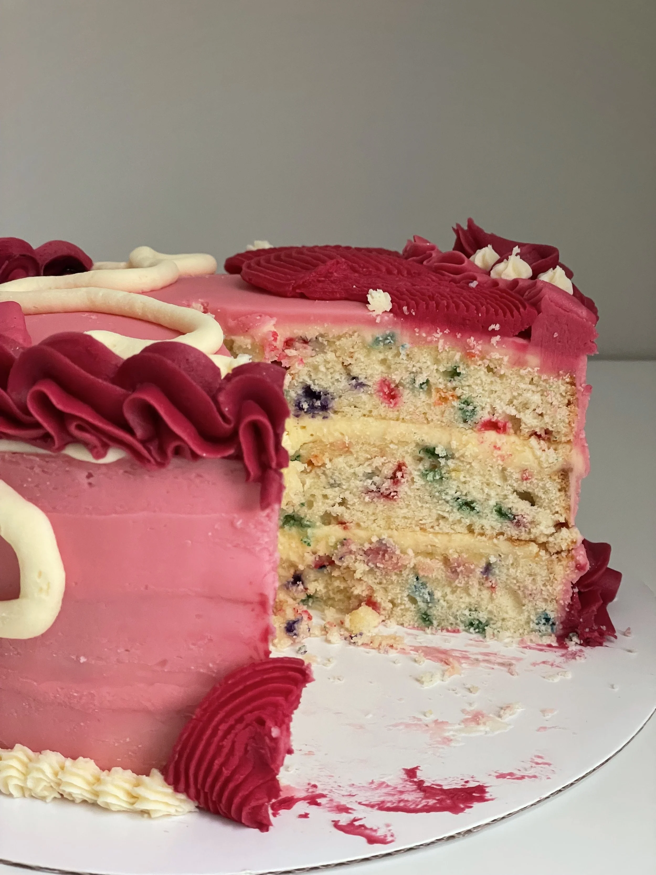 A slice of pink birthday cake with multicolored sprinkles inside, decorated with red, white, and pink icing, and topped with decorative pink and white frosting accents.