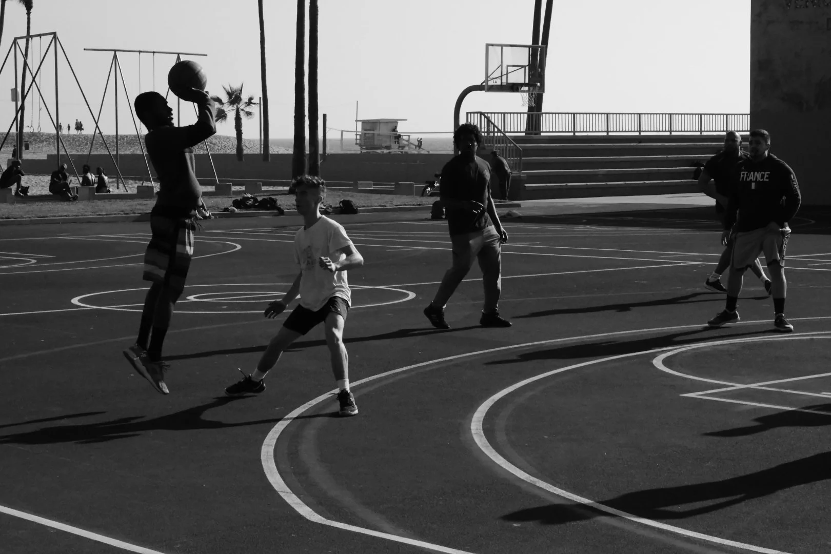 People playing basketball on an outdoor court near a beach with palm trees in the background during sunset.