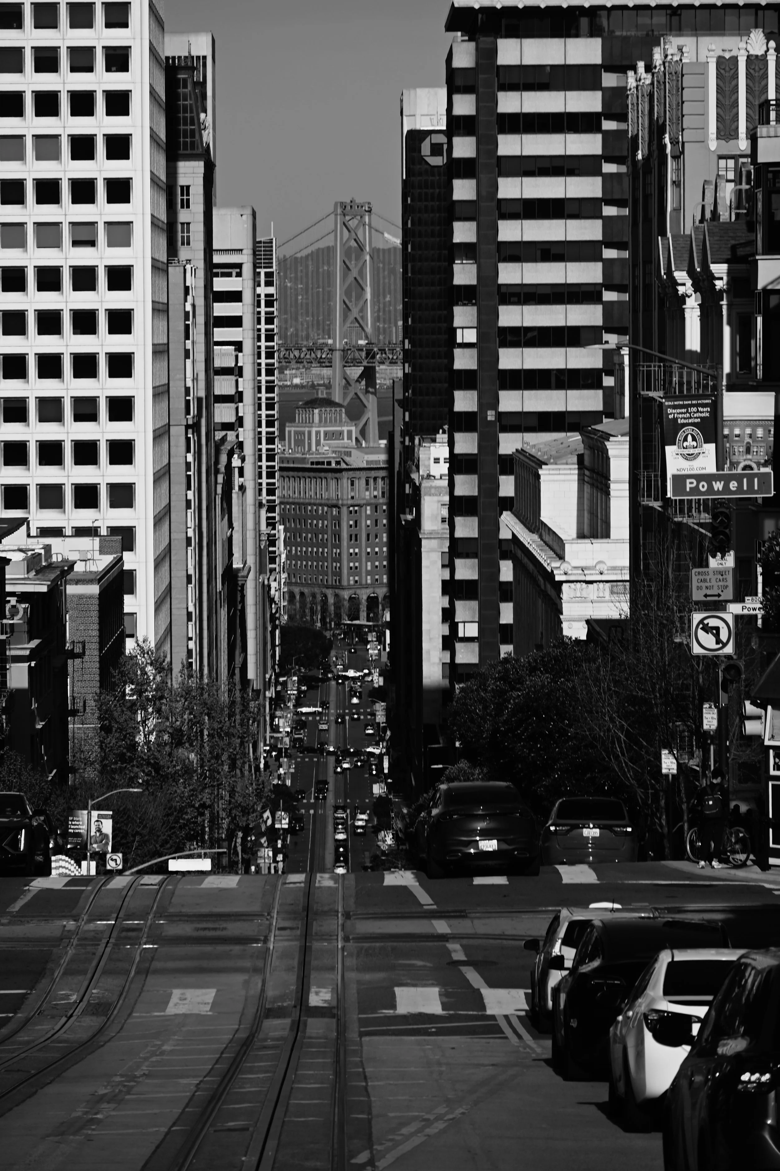 Black and white cityscape of tall buildings, billboards, and a crowded street with cars, and visible a bridge in the background.