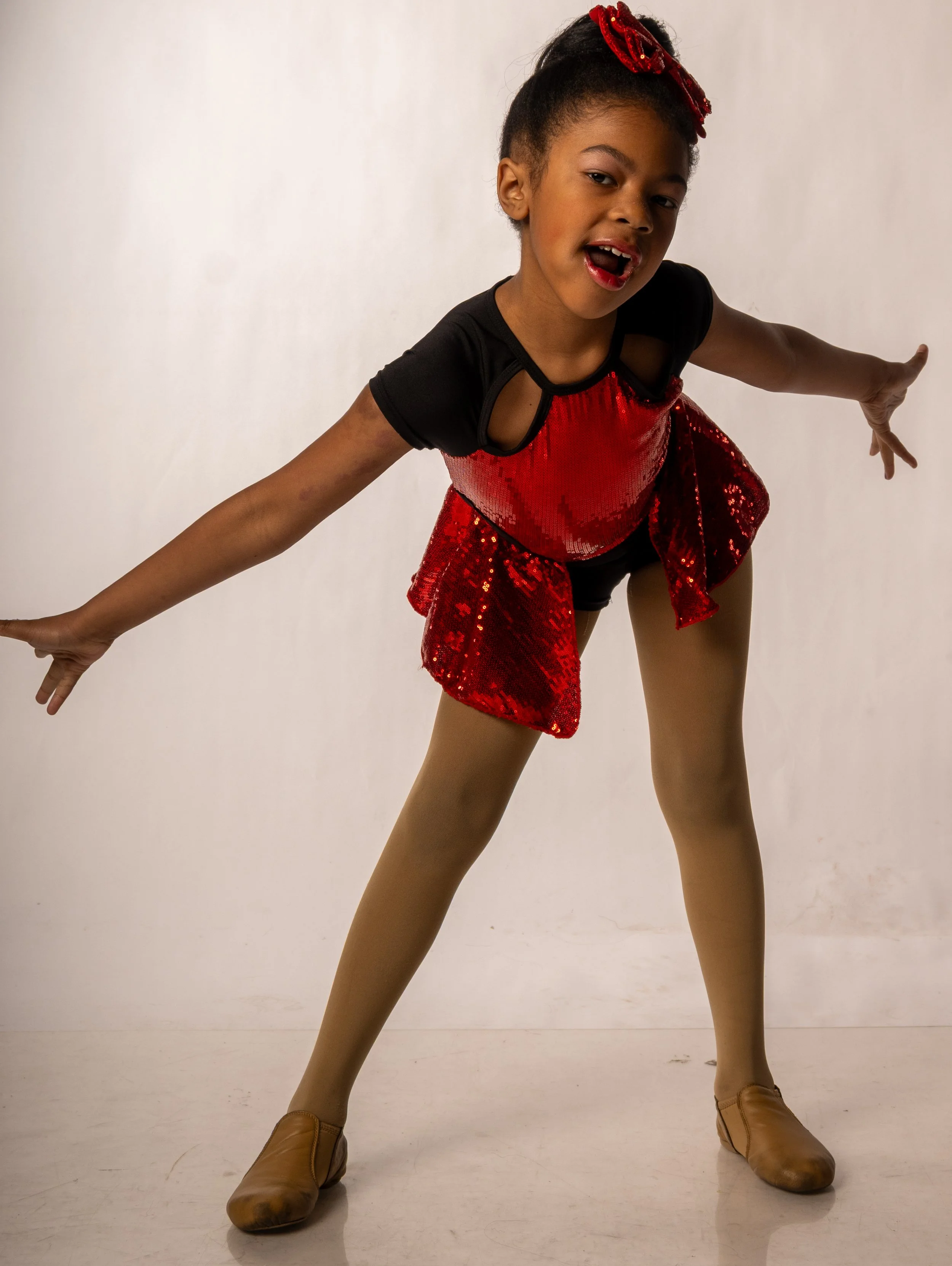 A young girl in a red sequined dance costume with tan tights and tan tap shoes, striking a dance pose with arms extended and legs apart.