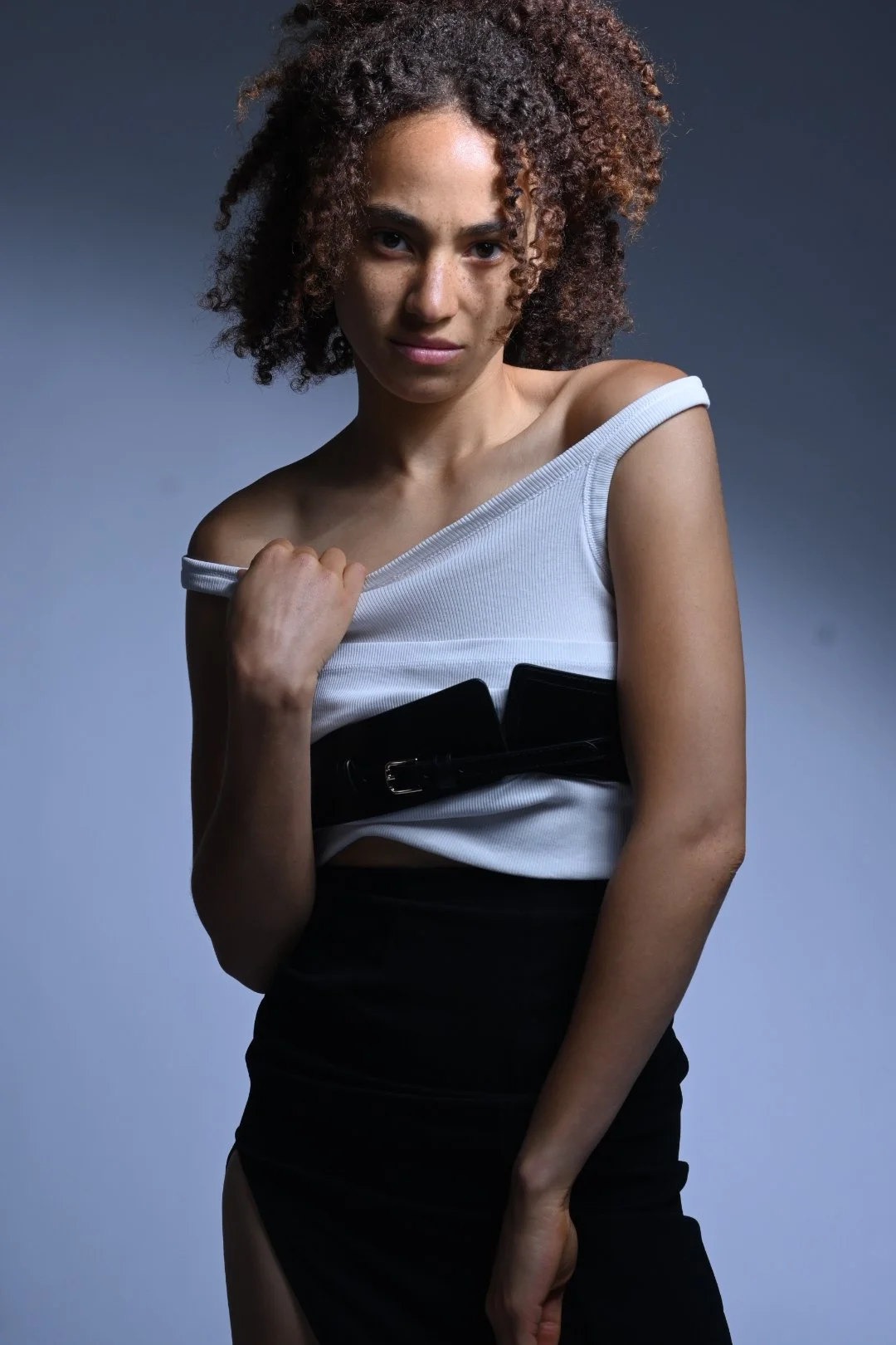A young woman with curly hair wearing a white top and black skirt with a large black belt, standing against a plain grey background.