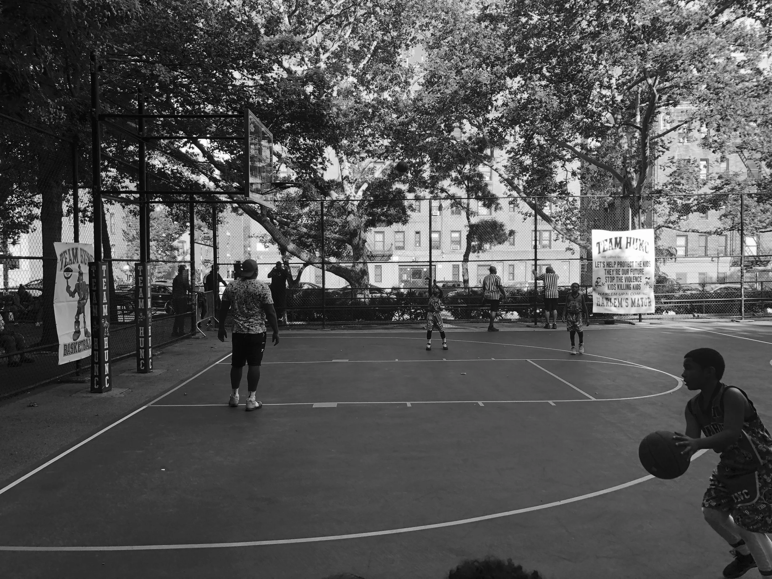 A black and white photo of children and adults playing basketball on an outdoor court surrounded by a fence and trees. There are posters on the fence and a sign that reads "TEAM HUNG" promoting protecting kids and stopping violence.
