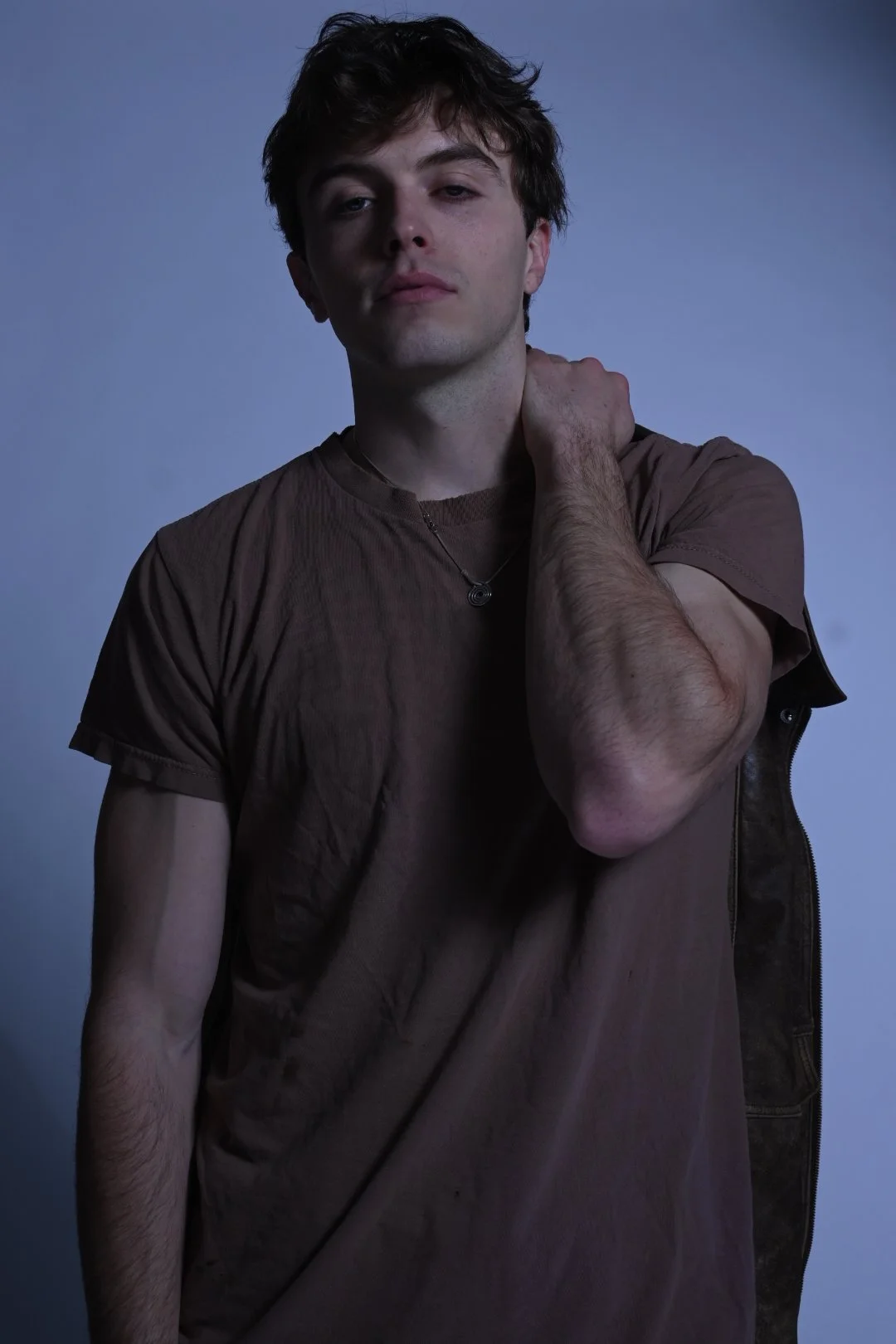 A young man with brown hair and a fair complexion is posing in front of a plain background, wearing a brown t-shirt and a necklace, with his hand on his neck, looking at the camera with a relaxed expression.