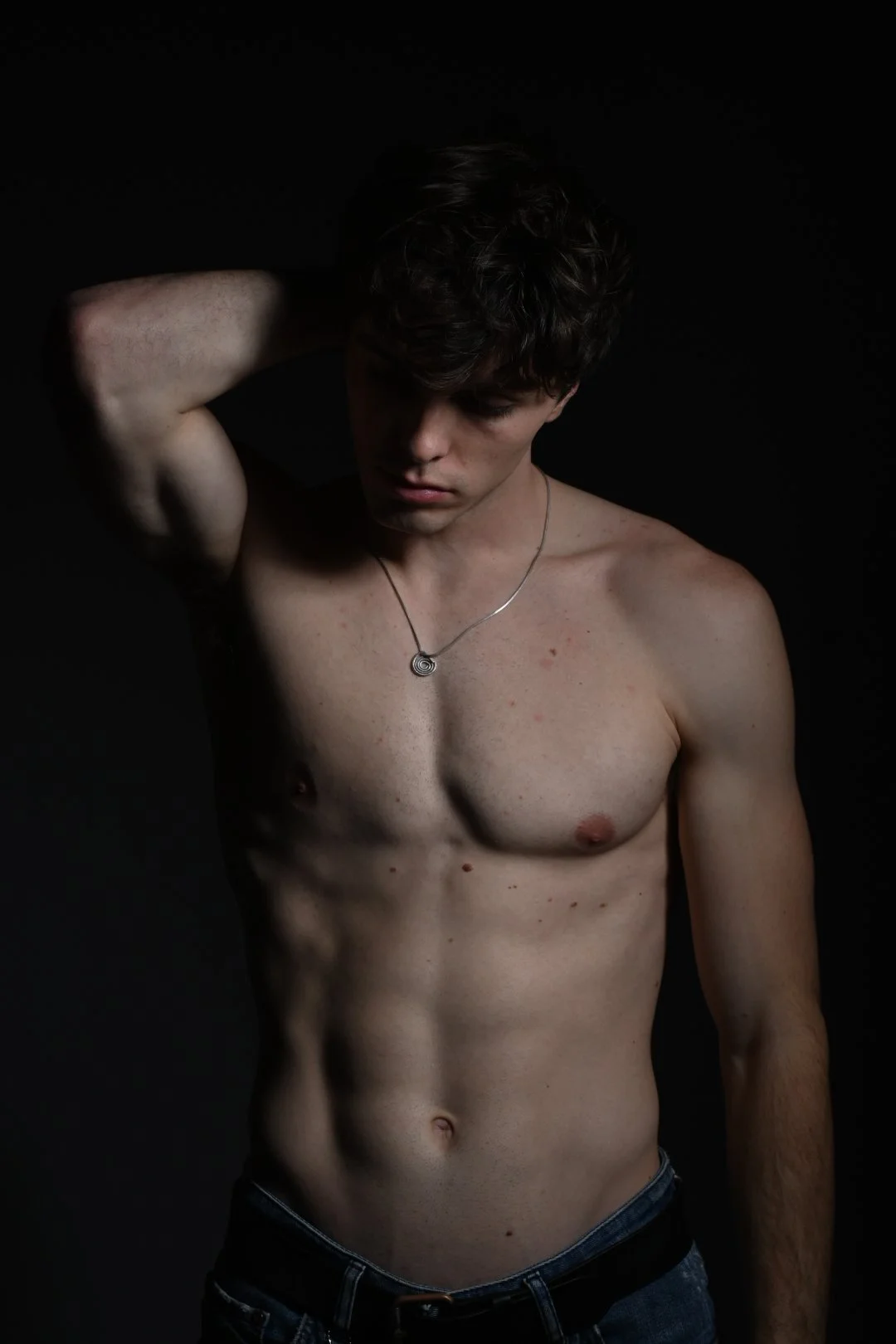 A shirtless young man with curly hair wearing a silver necklace, posing with one arm behind his head against a dark background.