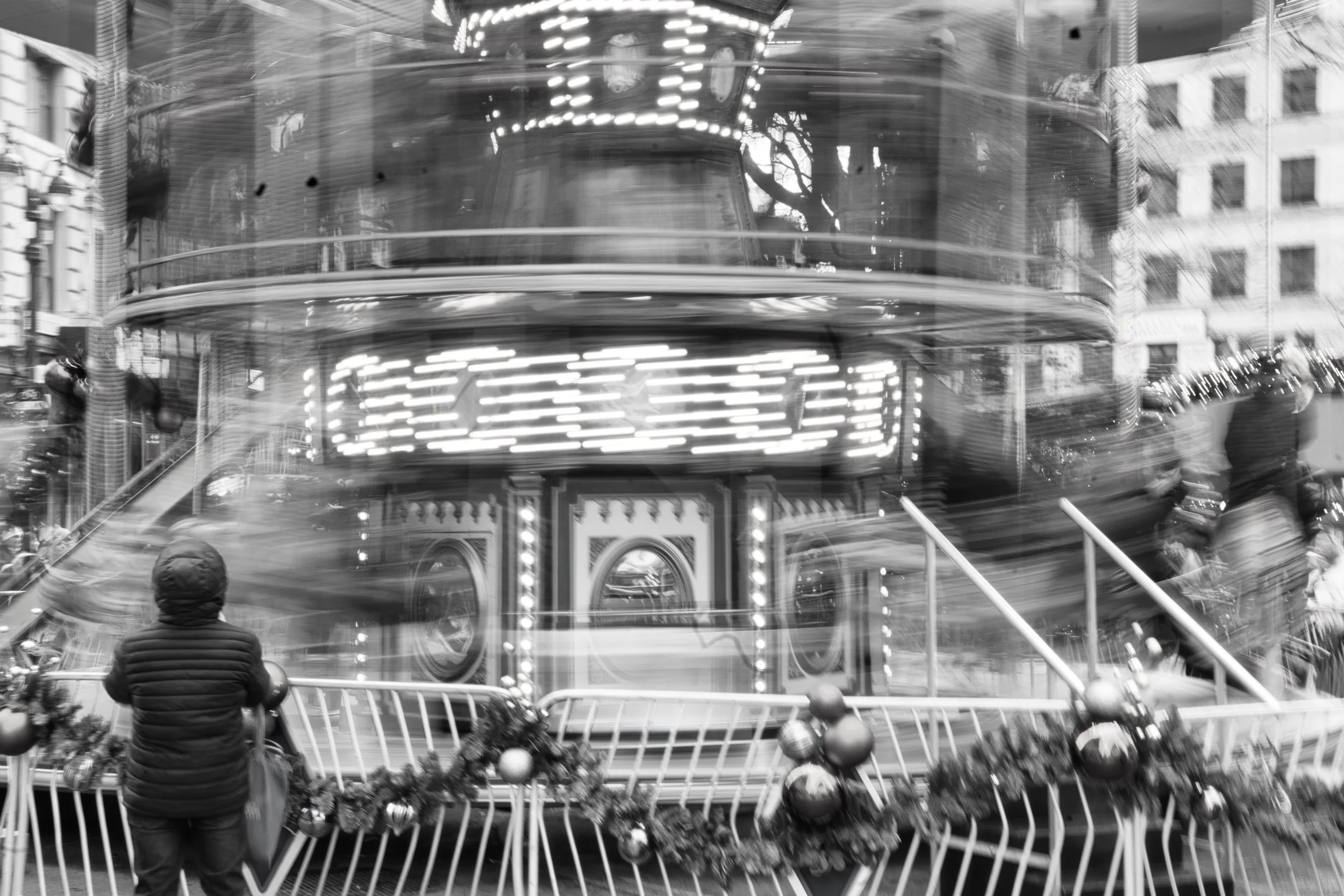 Black and white photo of a carousel with bright lights, a building, and people standing around it in an urban setting.