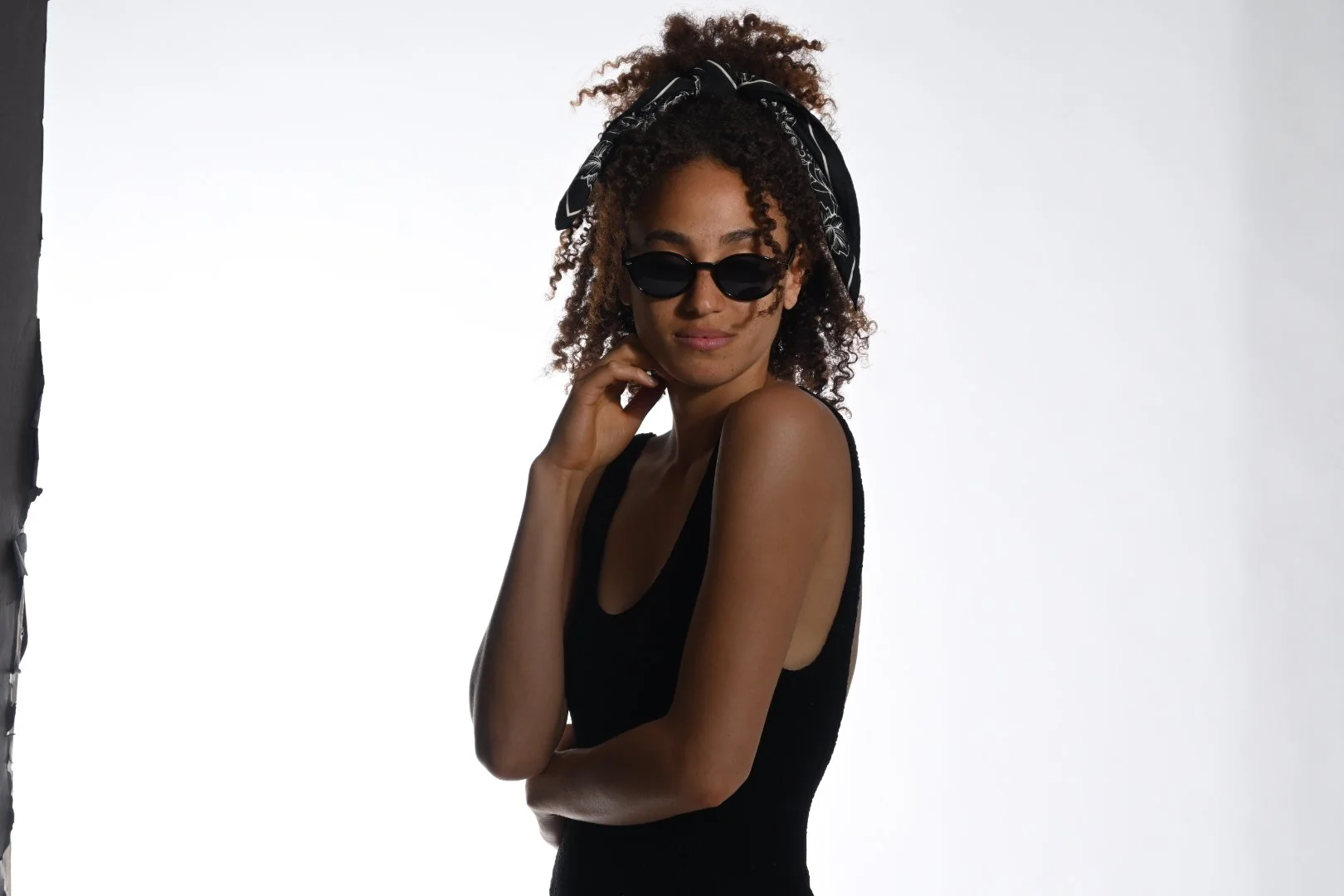 A woman with curly hair wearing black sunglasses, a black sleeveless top, and a black headscarf with white patterns, standing against a white background with a partial black backdrop on the left.