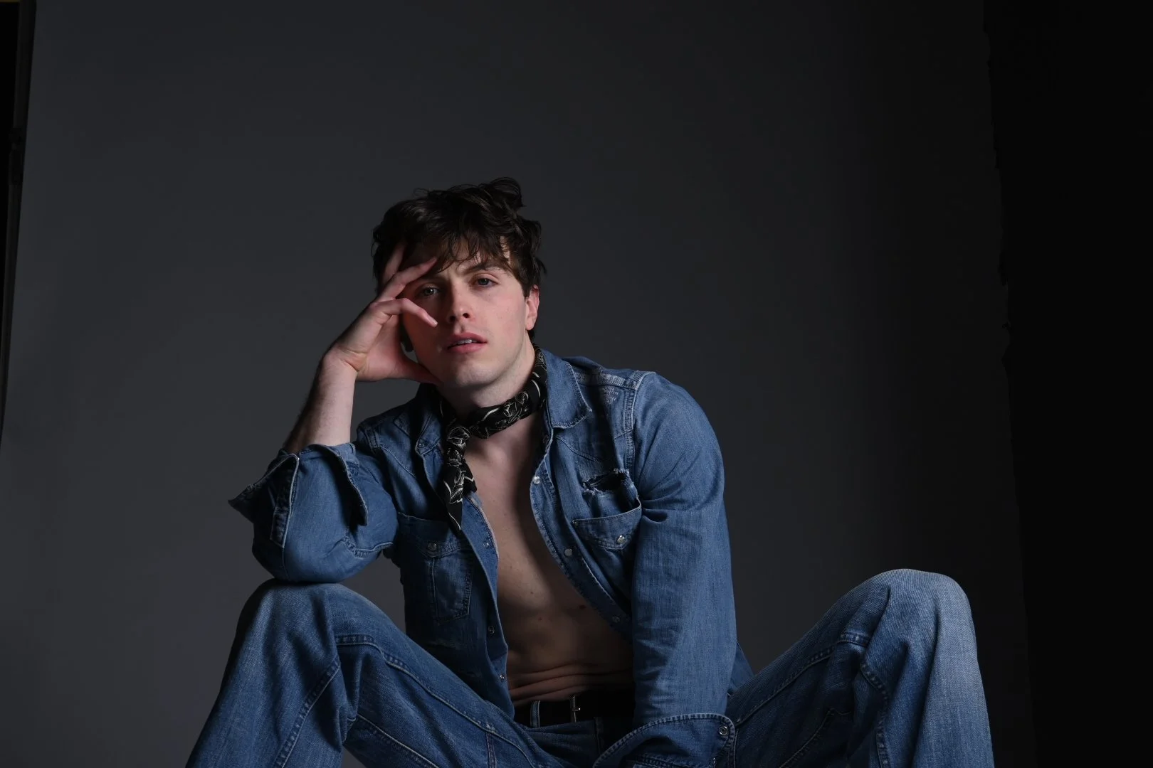 A young man with dark hair, wearing a denim jacket and jeans, sitting against a dark background. He has a black bandana around his neck and is posing with his hand on his face, gazing at the camera.