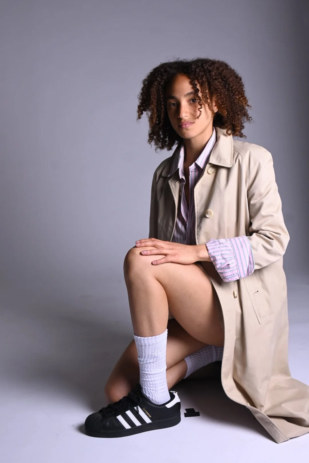 A young woman with curly hair kneeling, wearing a beige trench coat, striped shirt, white socks, and black Adidas sneakers, against a plain gray background.