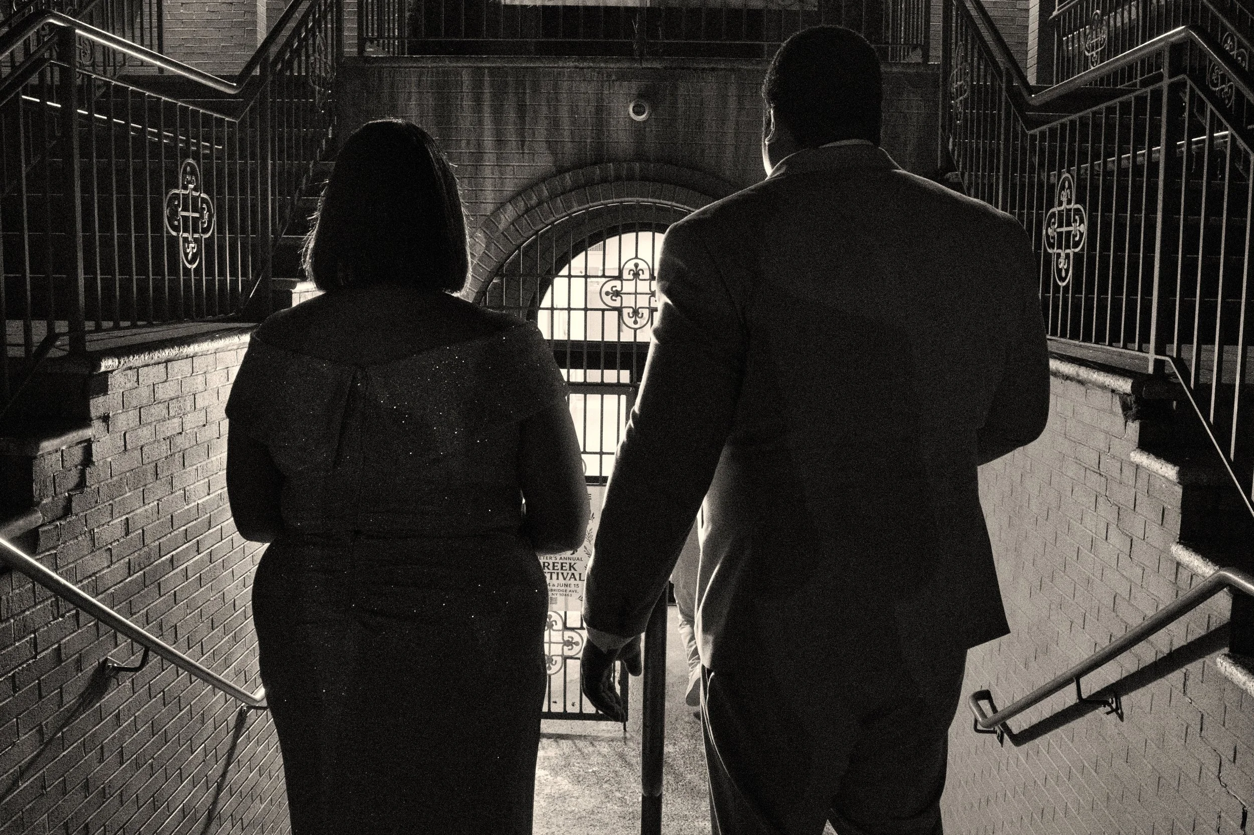 Two people holding hands and walking up stairs towards an arched gate, black and white photo.