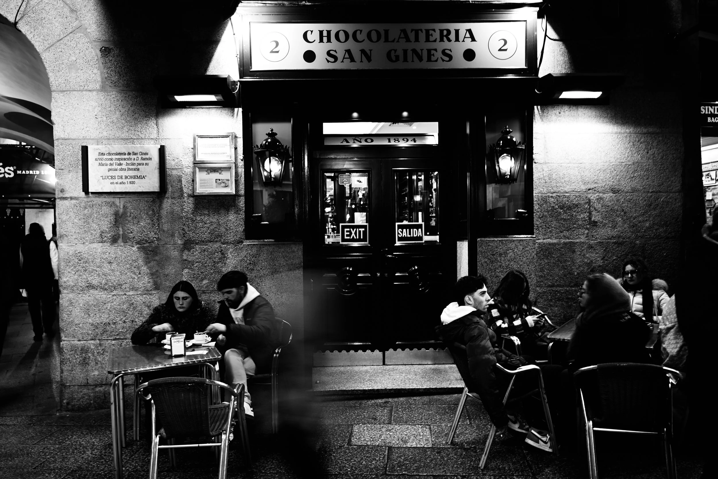 A black and white photo of the entrance to a chocolate shop called "Chocolateria San Gines" with people sitting at tables outside under streetlights.