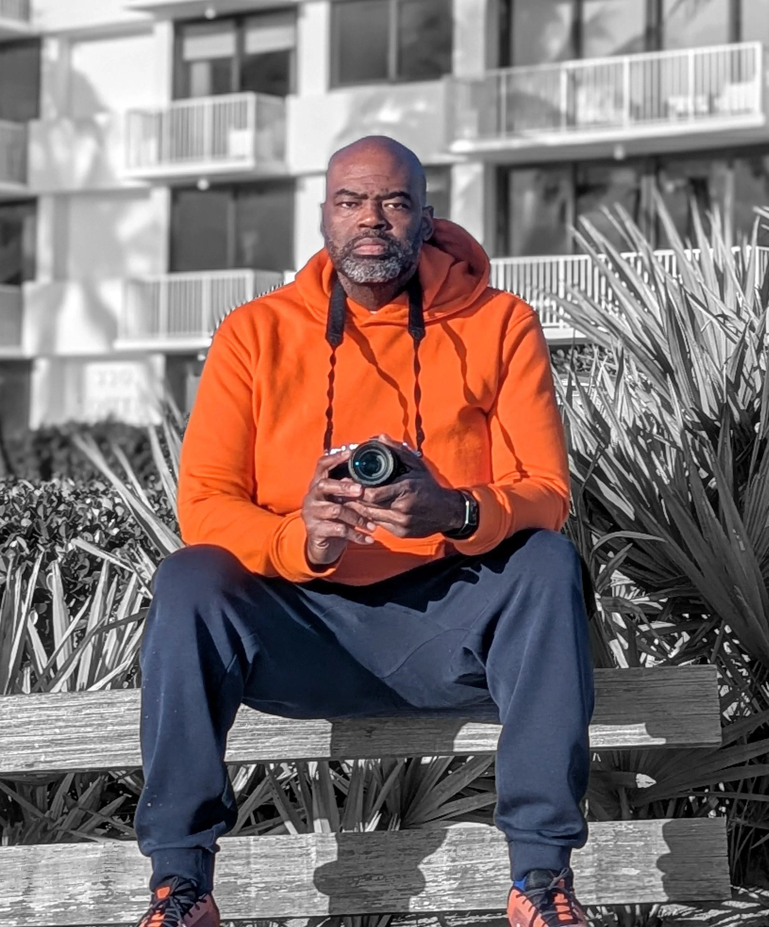 A man sitting on a bench holding a camera, wearing an orange hoodie and black pants, with a modern apartment building and plants behind him.
