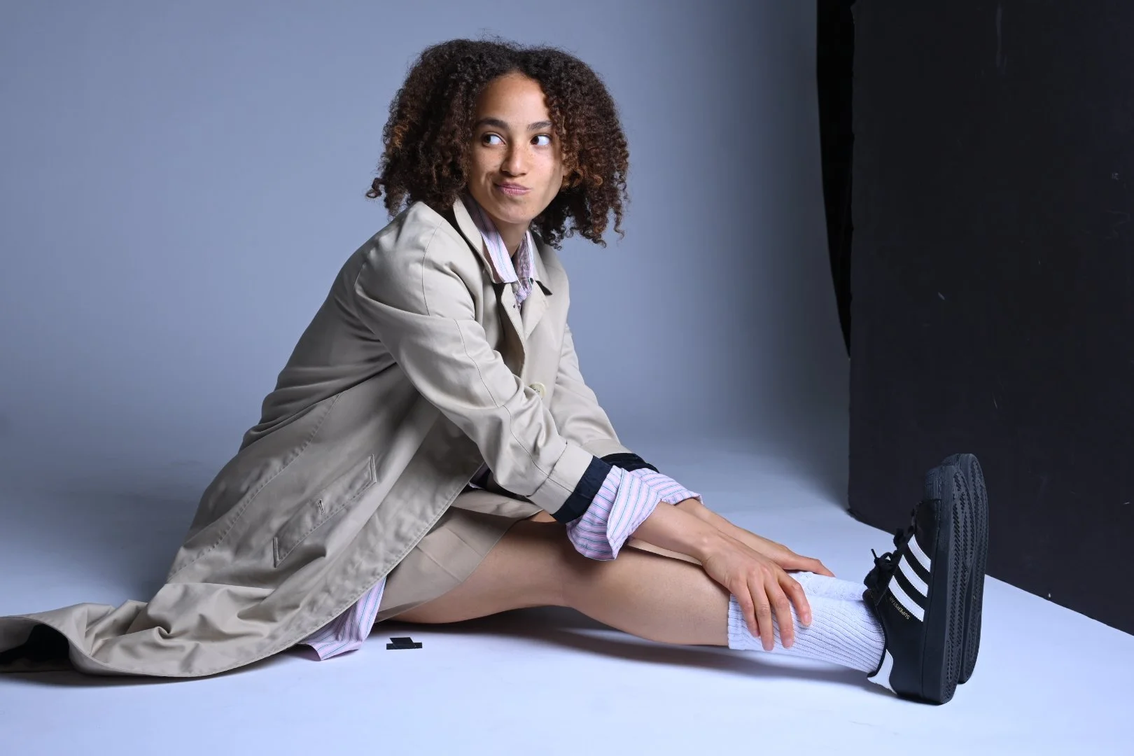 A person with curly hair wearing a beige coat, white shorts, and black Adidas sneakers, sitting on the floor and stretching their leg.
