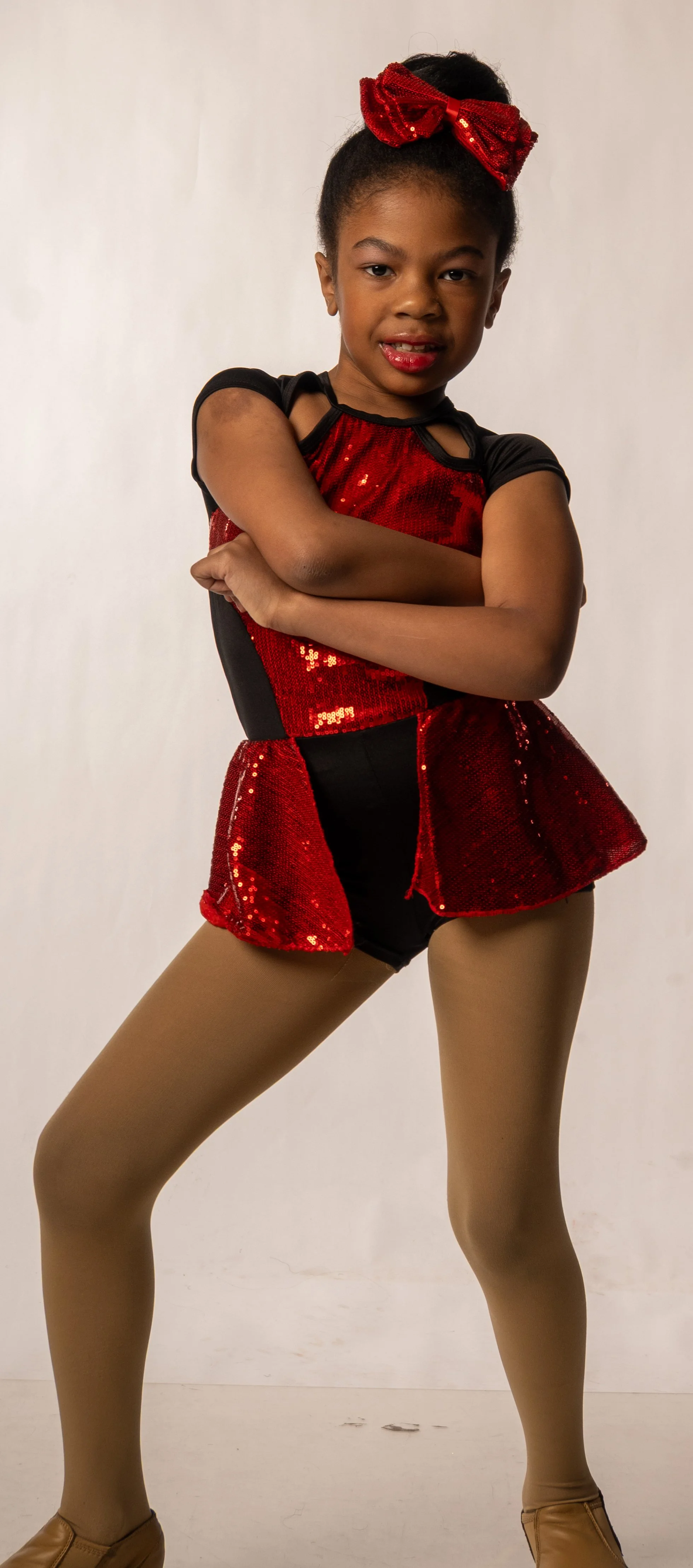 Young girl dressed in a red and black dance costume with a large red sequin bow in her hair, posing with arms crossed on a plain white background.