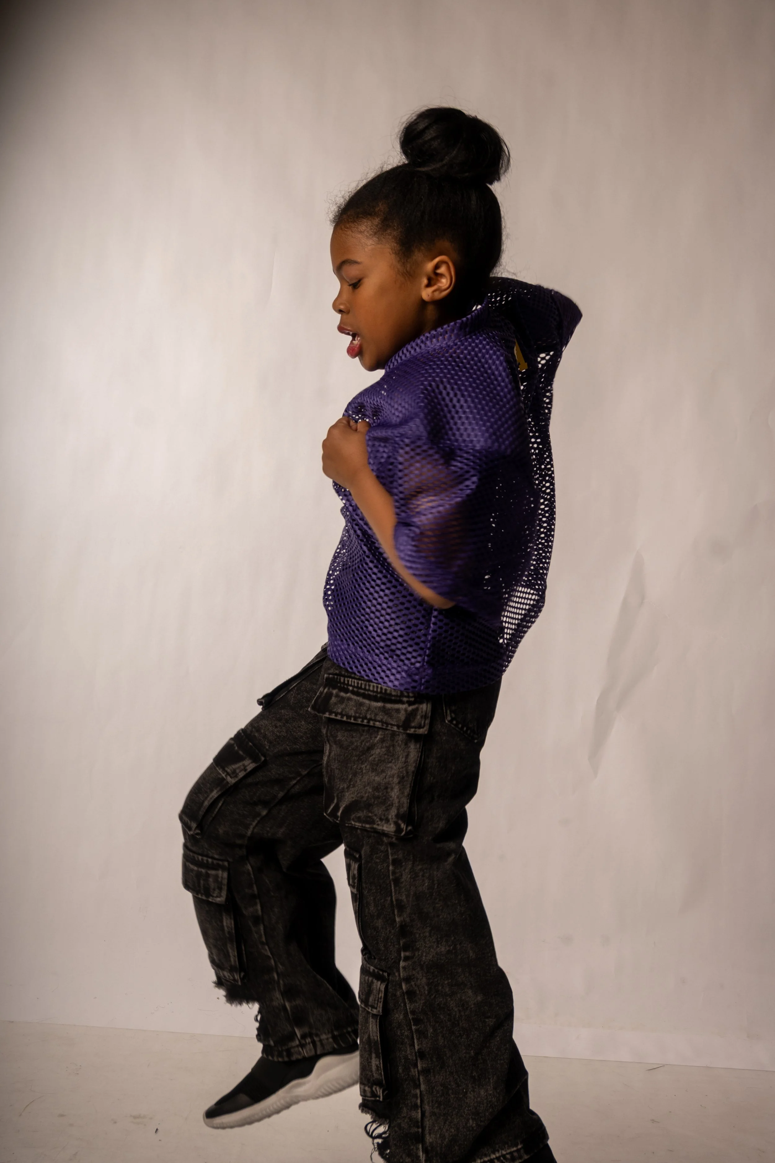 A young girl with dark hair in a bun, wearing a purple mesh hoodie, black cargo pants, and sneakers, is standing against a plain white background. She is looking down with her eyes closed and mouth slightly open.