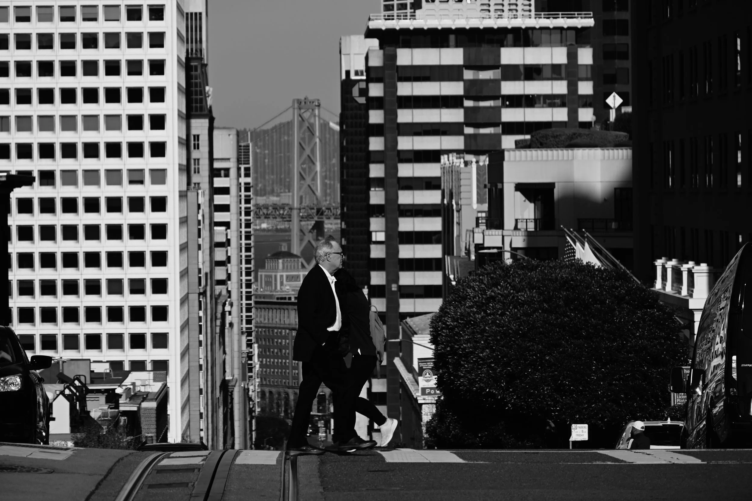 A black and white photo of a city street with tall buildings in the background. Three people are walking and talking in the foreground, with one of them smoking a cigarette. There are parked cars and some trees on the street.
