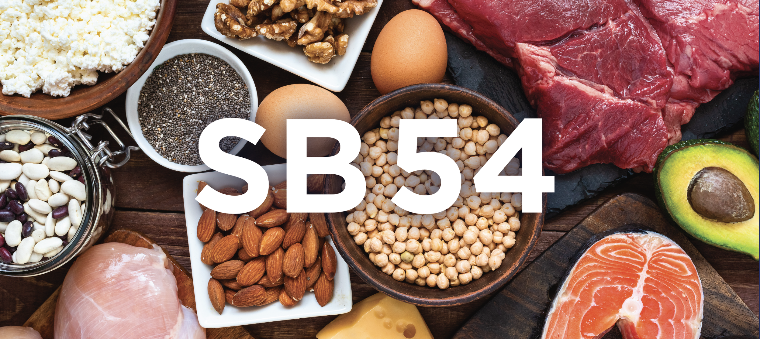 A variety of fresh ingredients including eggs, nuts, seeds, cheese, chicken, salmon, avocado, and beef arranged on a wooden surface, with a