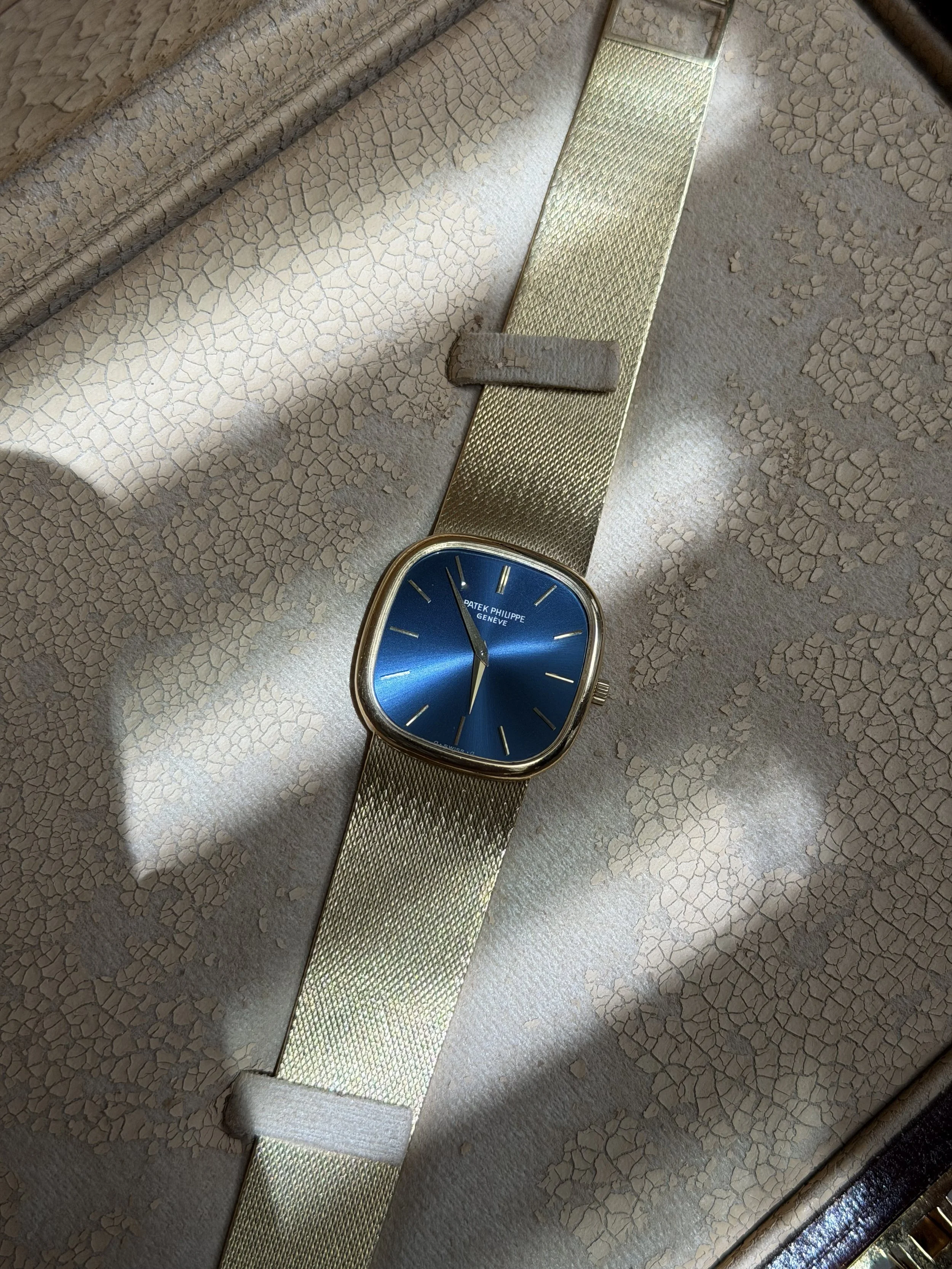 A gold wristwatch with a blue face and gold strap, placed on a cracked ceramic surface with a textured gold slot underneath.