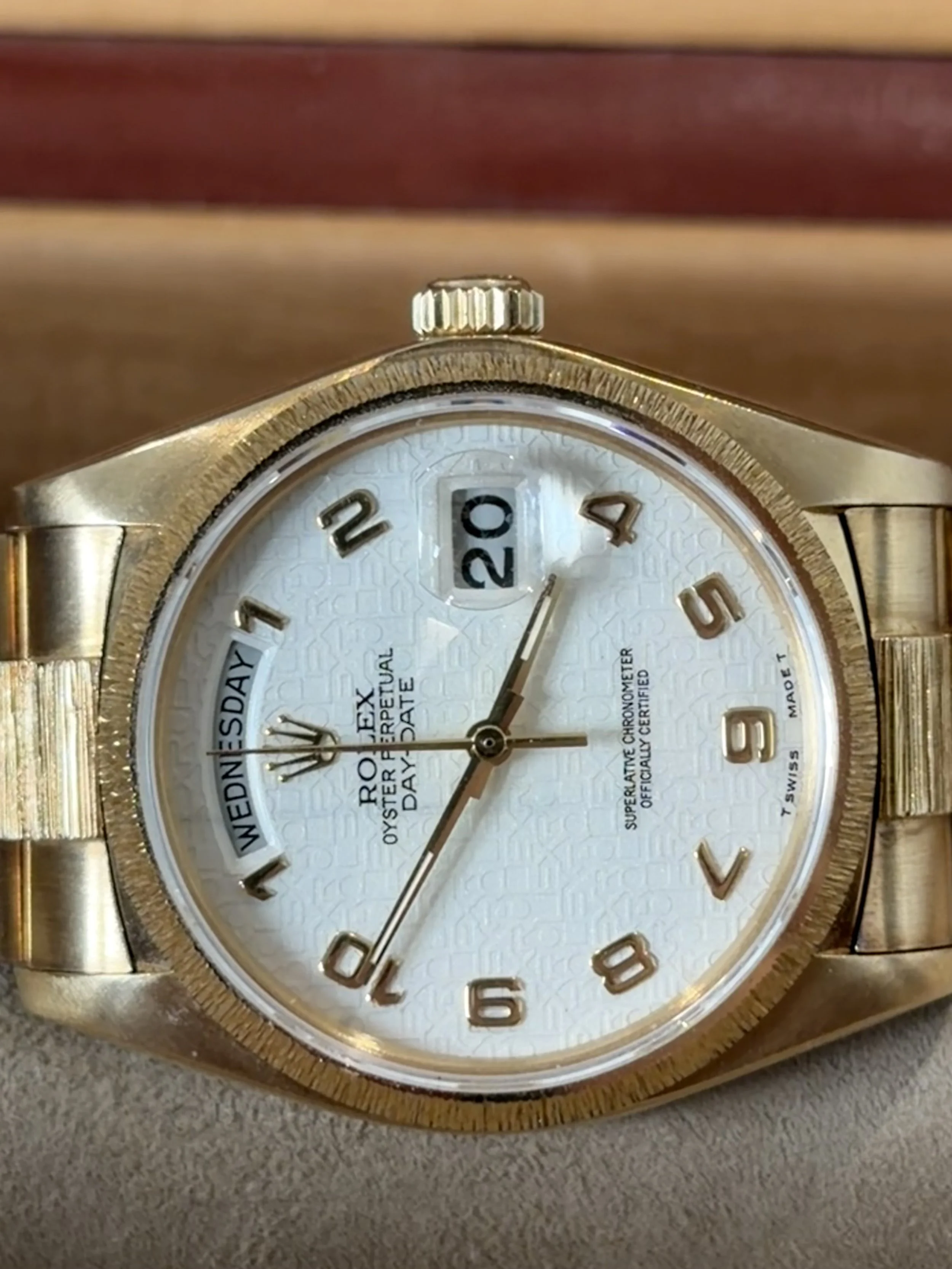 Close-up of a gold wristwatch with a white textured dial, gold numerals, and a date window at 3 o'clock, resting on a beige surface.