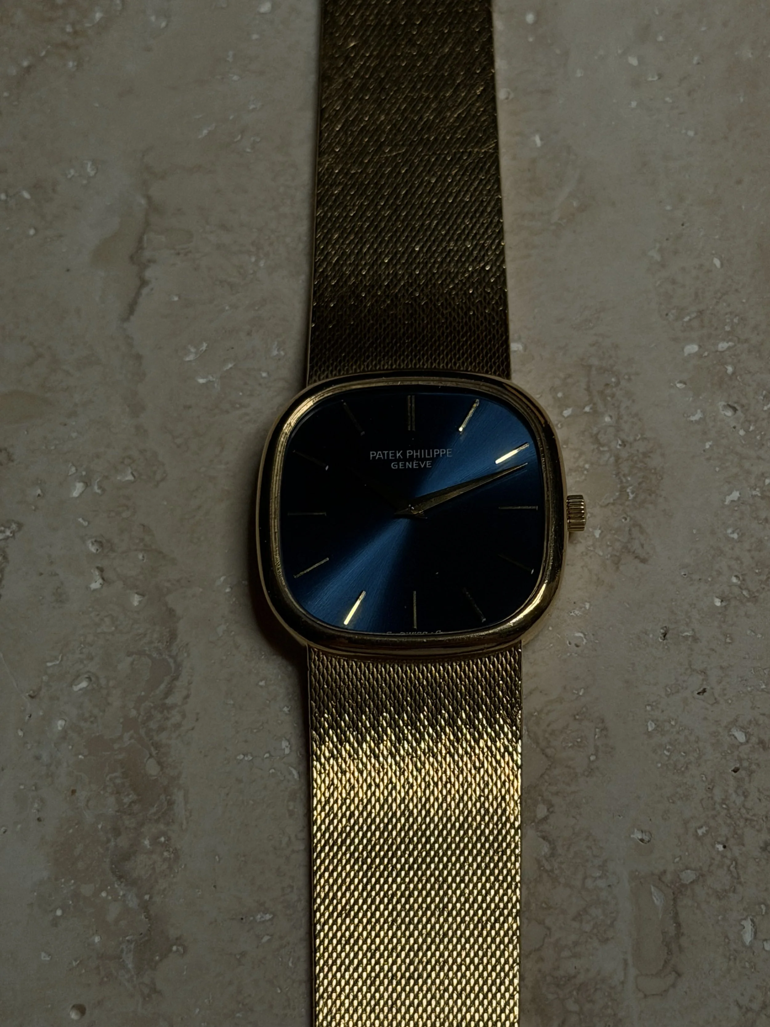 A gold wristwatch with a black dial face and gold markers, featuring the words "Patek Philippe Genève" on the face, resting on a textured, light-colored stone surface.